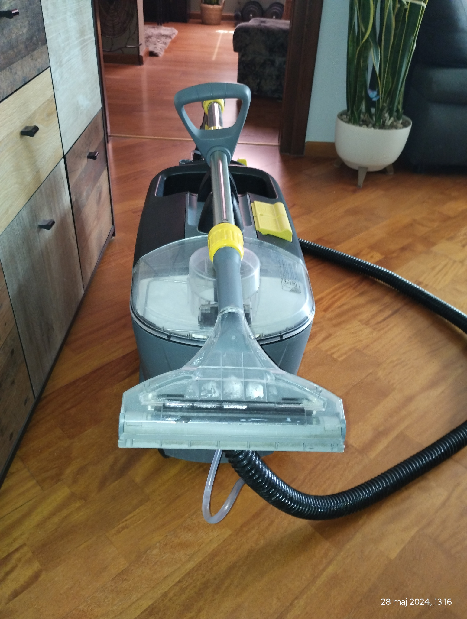 Urządzenie do czyszczenia dywanów i tapicerki z przezroczystą końcówką na drewnianej podłodze, widoczny wąż i zbiornik na wodę.