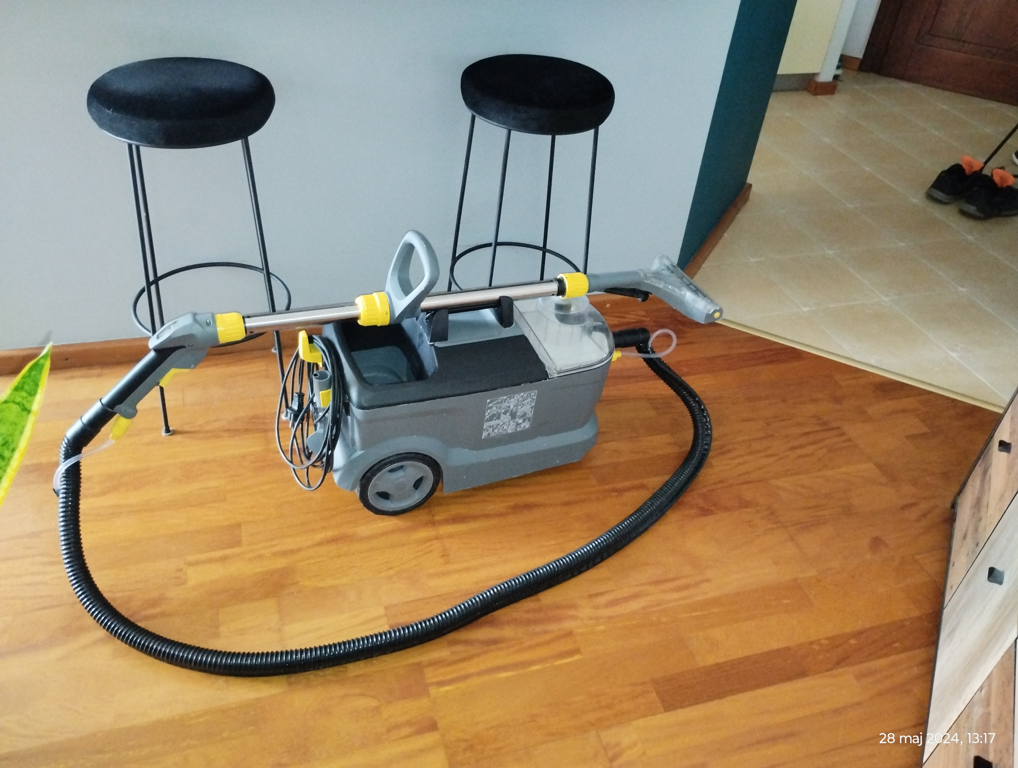 Profesjonalny odkurzacz piorący Karcher na drewnianej podłodze, z widocznymi dwoma czarnymi stołkami barowymi w tle, gotowy do użycia.