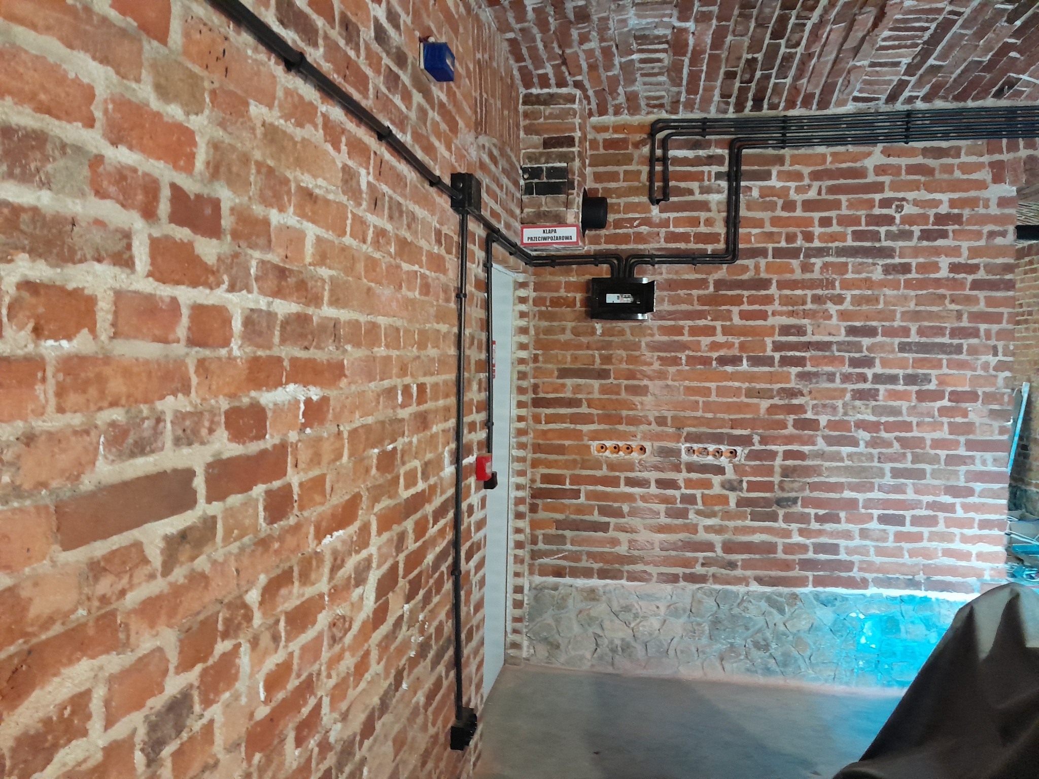 Instalacja elektryczna natynkowa w czarnych rurkach na ceglanej ścianie z widocznymi puszkami i gniazdkami. Widoczny fragment klapy przeciwpożarowej.