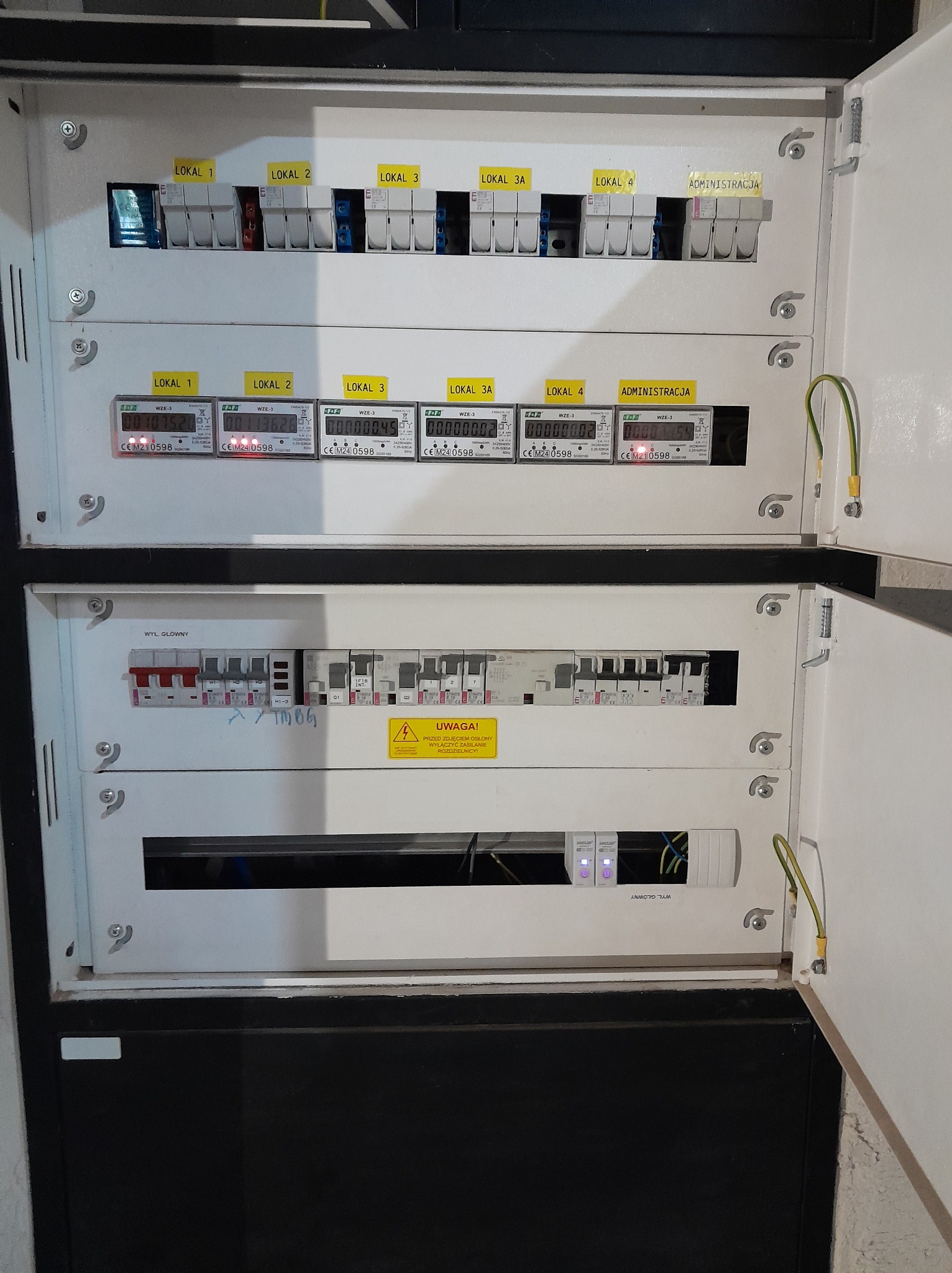 Rozdzielnica elektryczna z licznikami energii dla lokali i administracji. Widoczne bezpieczniki, wyłączniki i przewody. Otwarta obudowa rozdzielnicy.