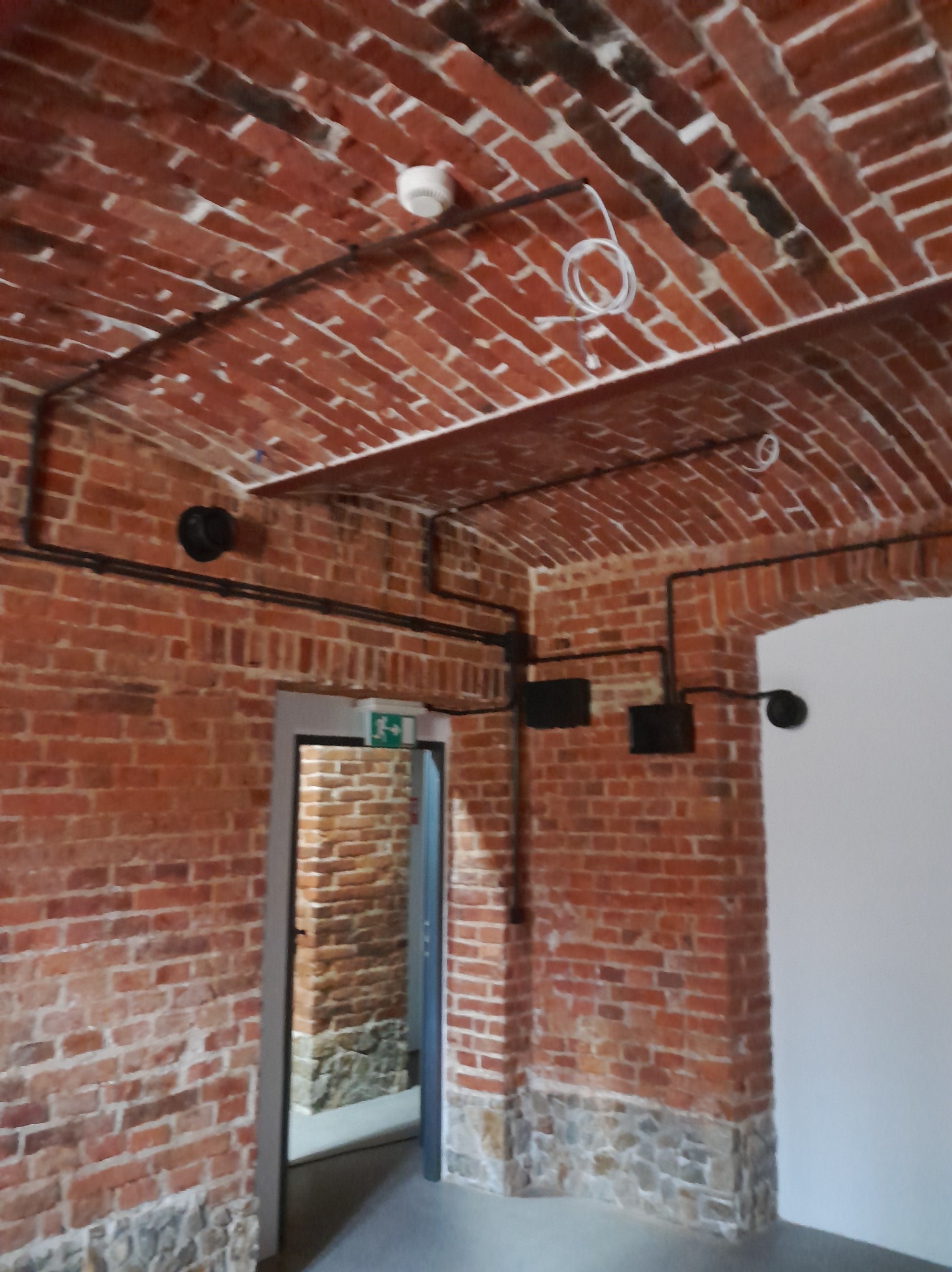 Instalacja elektryczna natynkowa w ceglanym wnętrzu z łukowym sufitem. Widoczne czarne rury, puszki i przewody. Biały czujnik dymu na suficie. Surowy, industrialny styl.