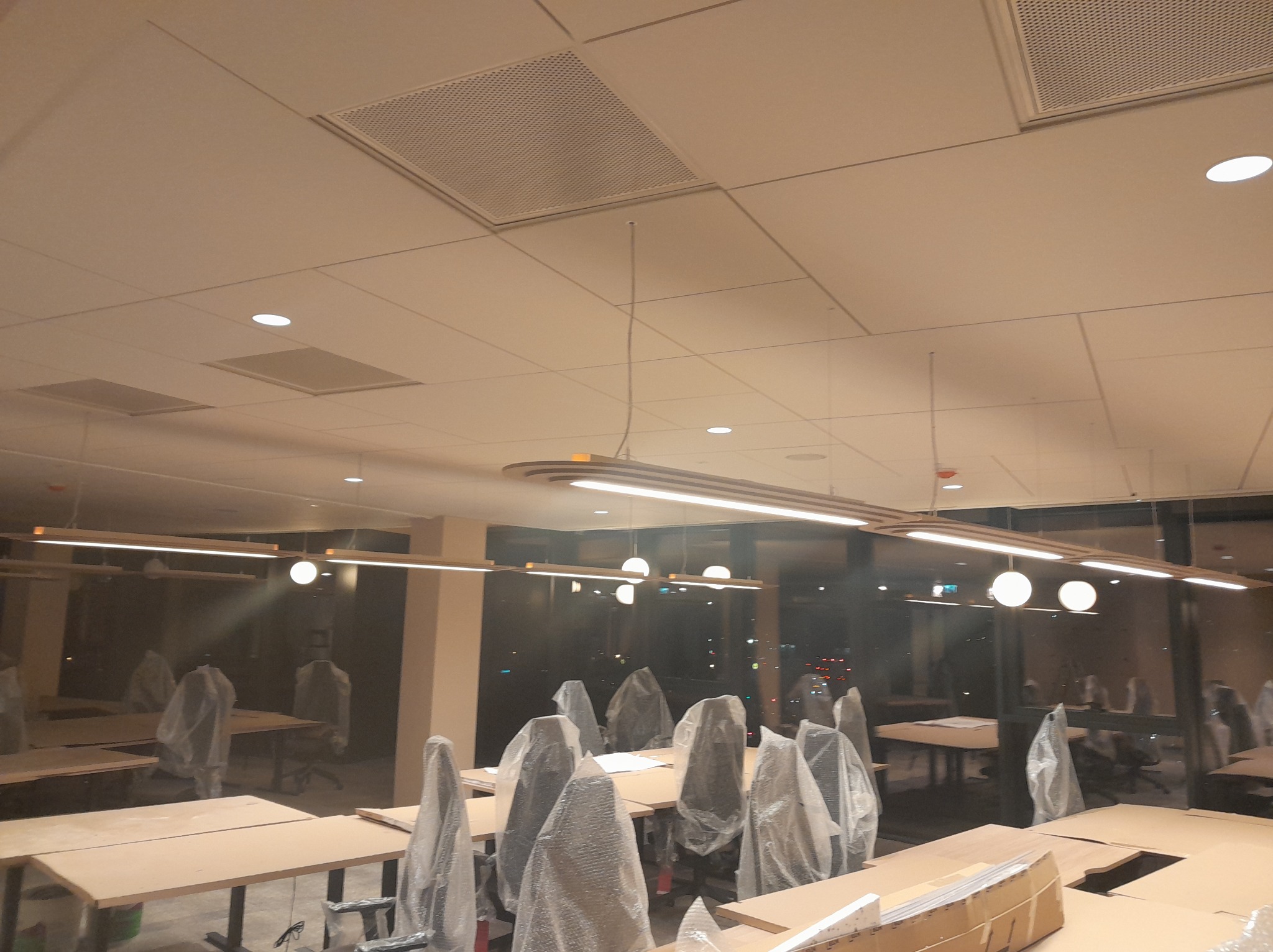 Biuro w trakcie wykańczania: widoczne lampy liniowe LED pod sufitem, biurka z krzesłami w folii ochronnej. Oświetlenie biurowe w nowej przestrzeni.