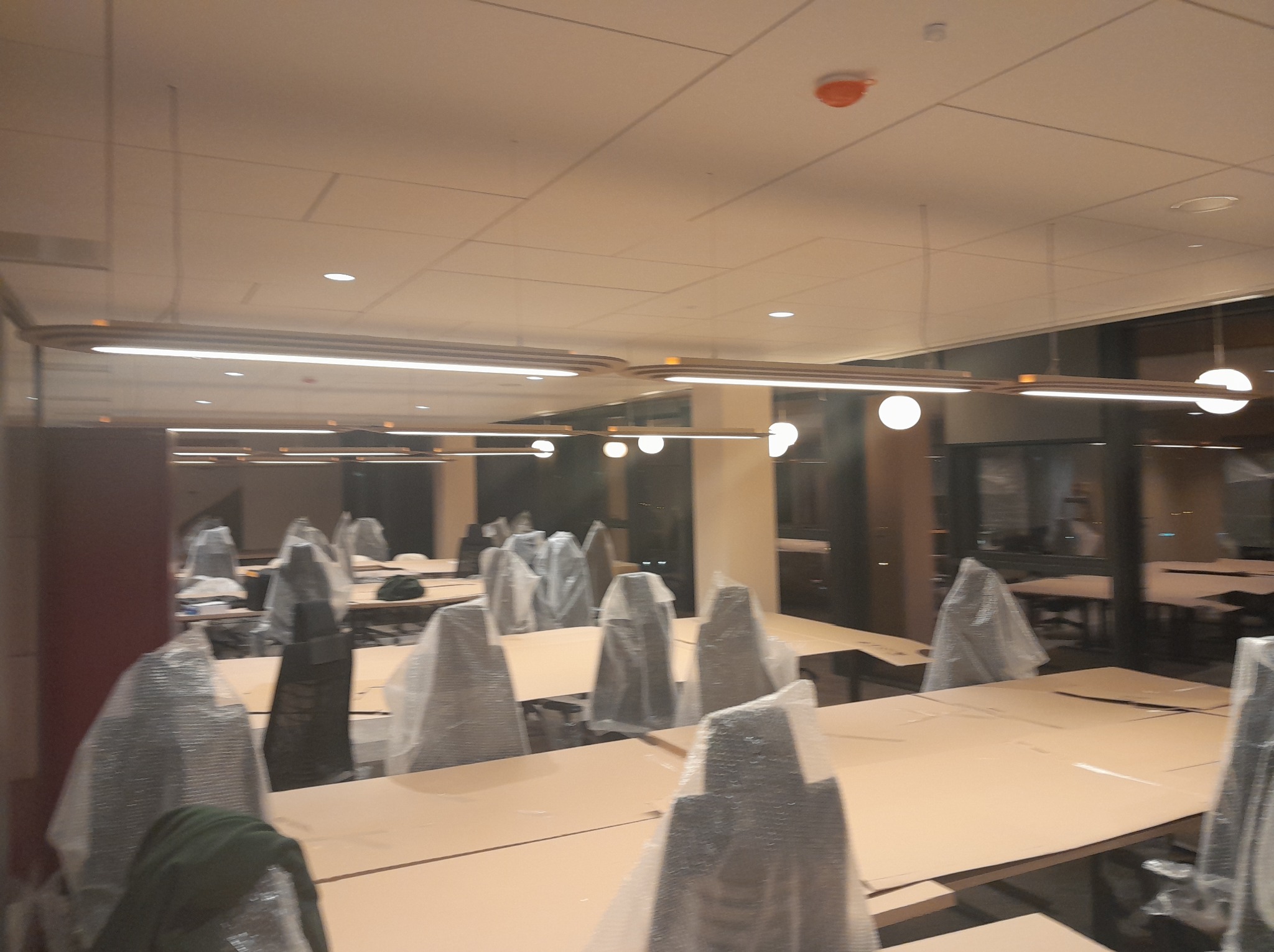 Biuro po remoncie z nowym oświetleniem: długie lampy LED nad stołami, krzesła w folii ochronnej. Widok na przestrzeń biurową z równomiernym, jasnym oświetleniem.