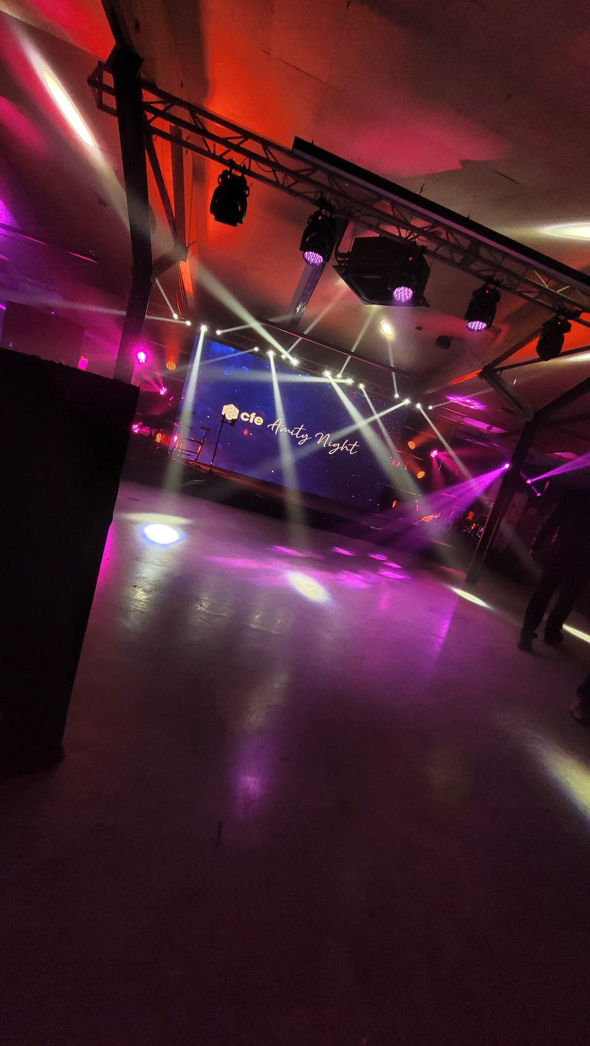 Oświetlona scena z ekranem wyświetlającym napis 'cfe Amity Night', reflektory na konstrukcji kratownicowej, fioletowe i białe światła sceniczne oświetlają parkiet.