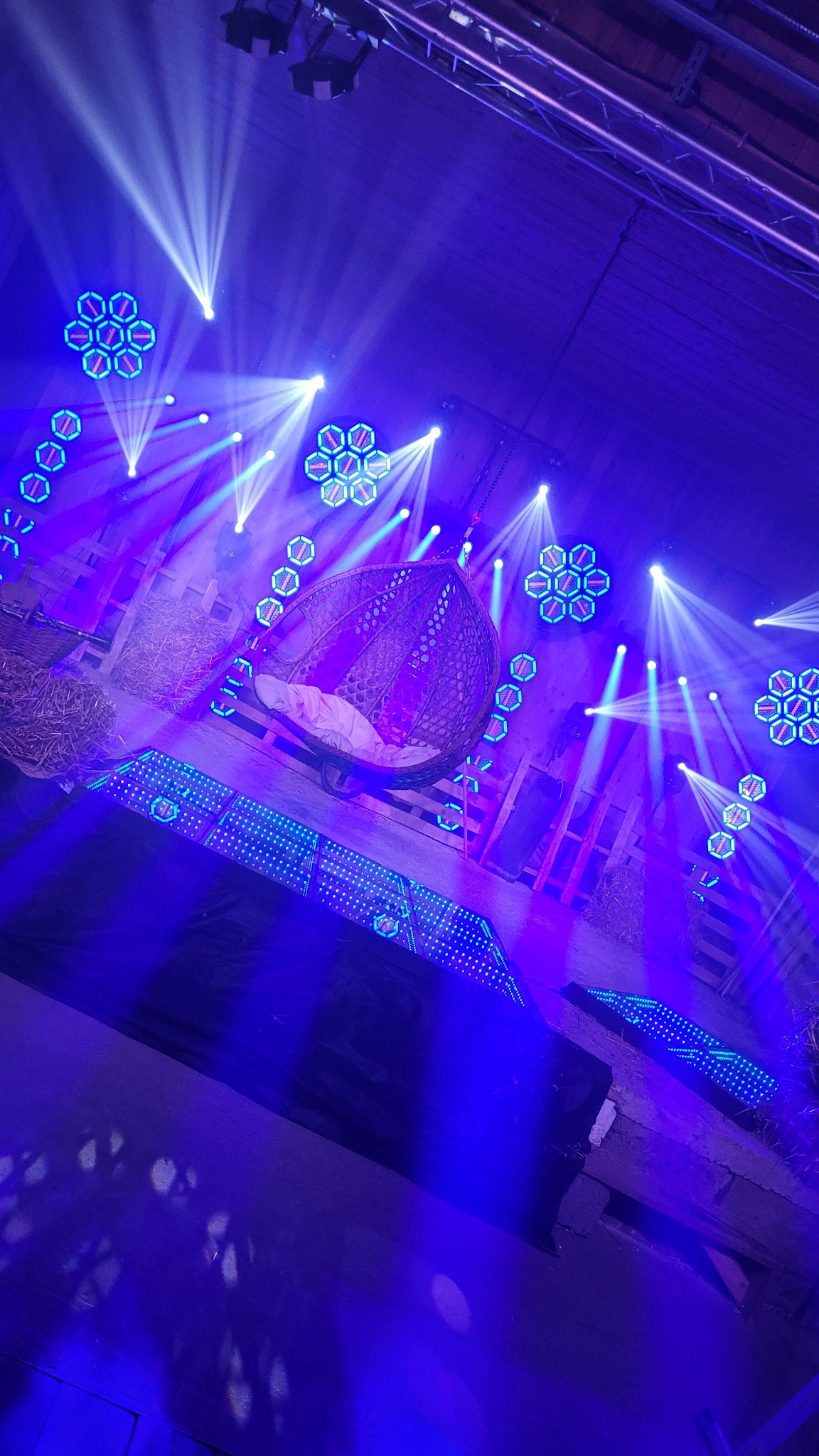 Scena oświetlona niebieskimi reflektorami, z wiszącym wiklinowym fotelem z białym kocem, na tle drewnianej konstrukcji i snopków siana.