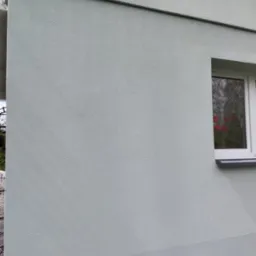 Jasnoszara elewacja budynku z widocznym oknem, na parapecie czerwone kwiaty w doniczce. Świeżo pomalowana powierzchnia z delikatną fakturą.