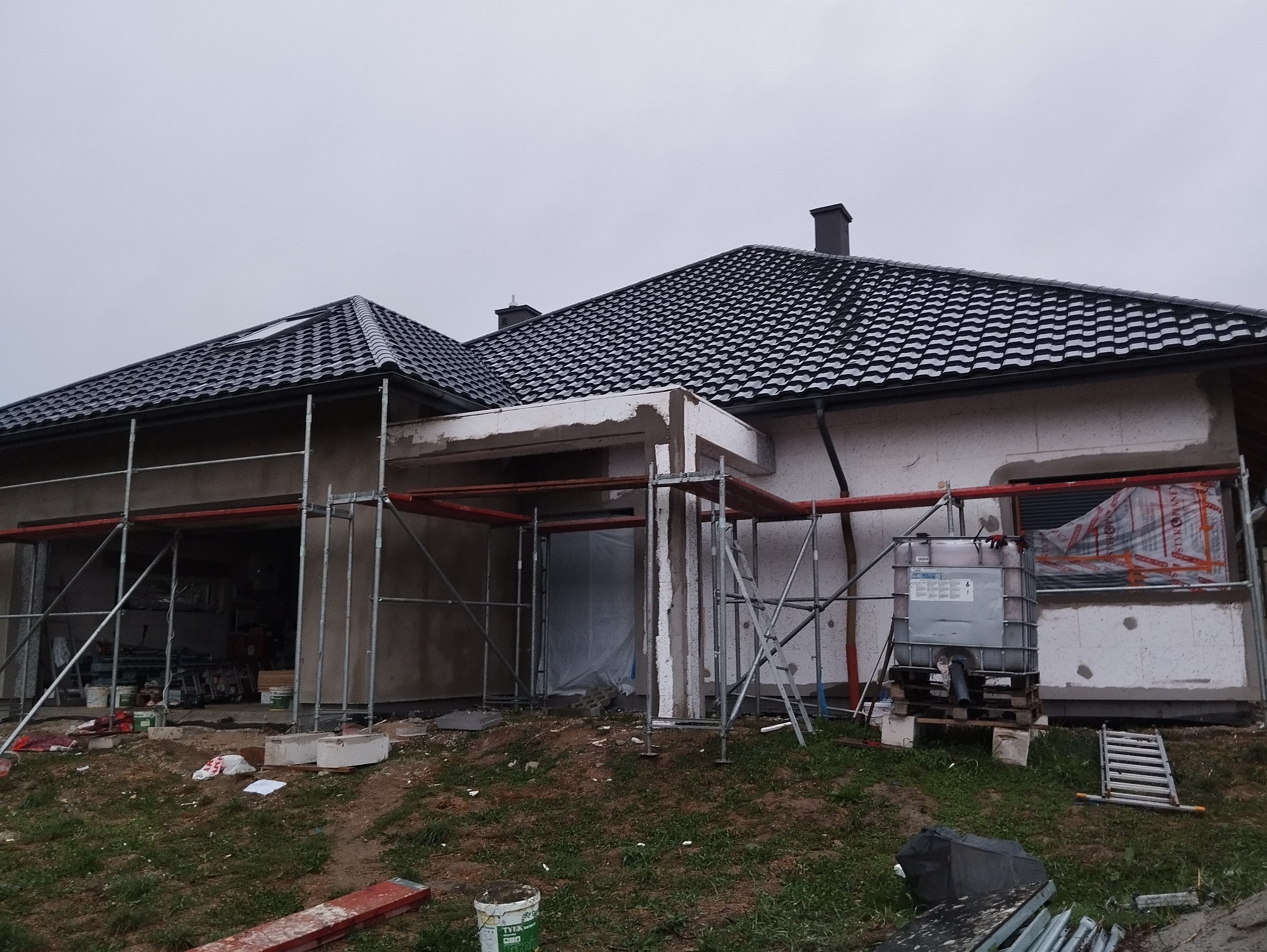 Dom w trakcie ocieplania elewacji, widoczne rusztowania, zbiornik na wodę, fragment ocieplonej ściany z siatką, oraz czarne dachówki na dachu.