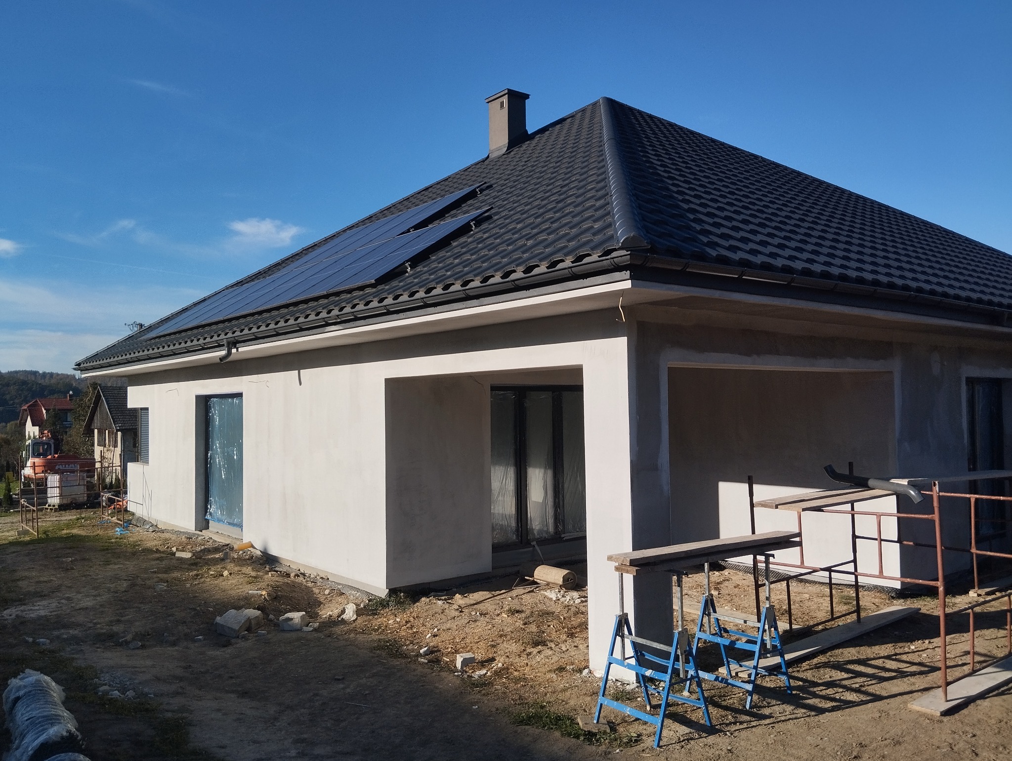 Surowy stan budynku jednorodzinnego z panelami słonecznymi na dachu, widoczne rusztowania i okna zabezpieczone folią.