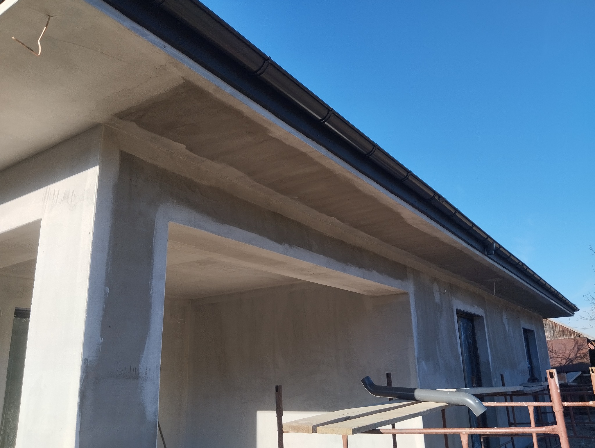 Surowa elewacja budynku w trakcie prac wykończeniowych, widoczne szare ściany, czarna rynna, błękitne niebo i fragment rusztowania.