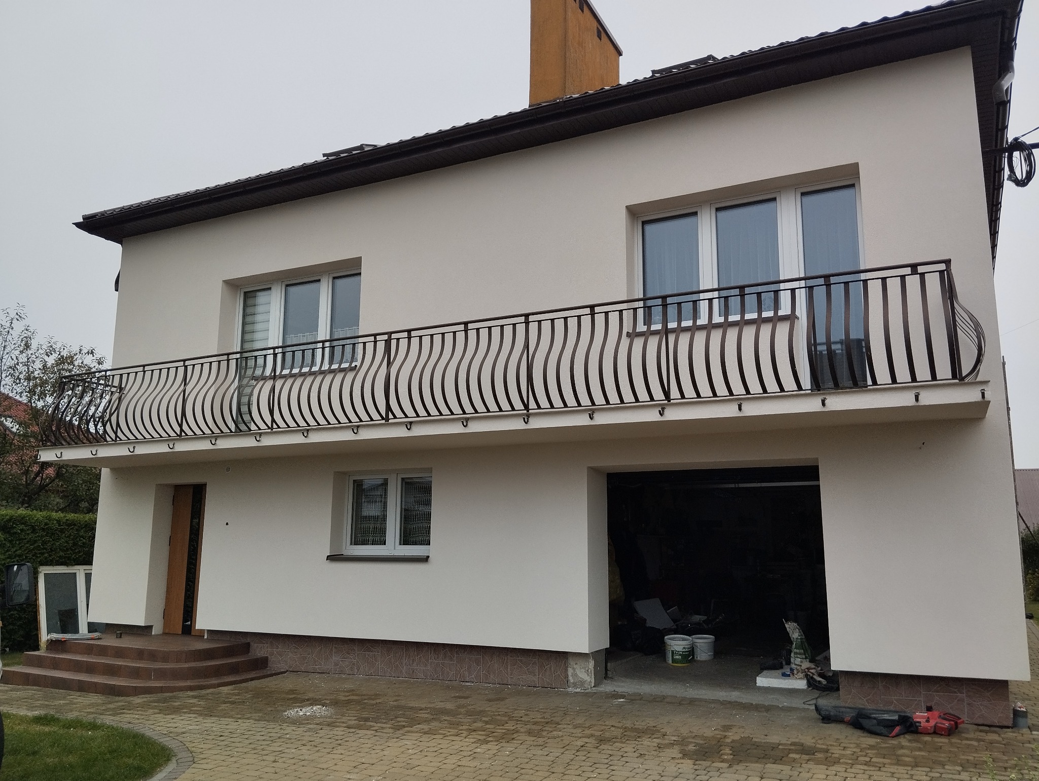 Dwupiętrowy dom z jasną elewacją, ciemnym dachem i balustradą balkonową o falistym wzorze; widoczne wejście do garażu z narzędziami i farbami w środku.