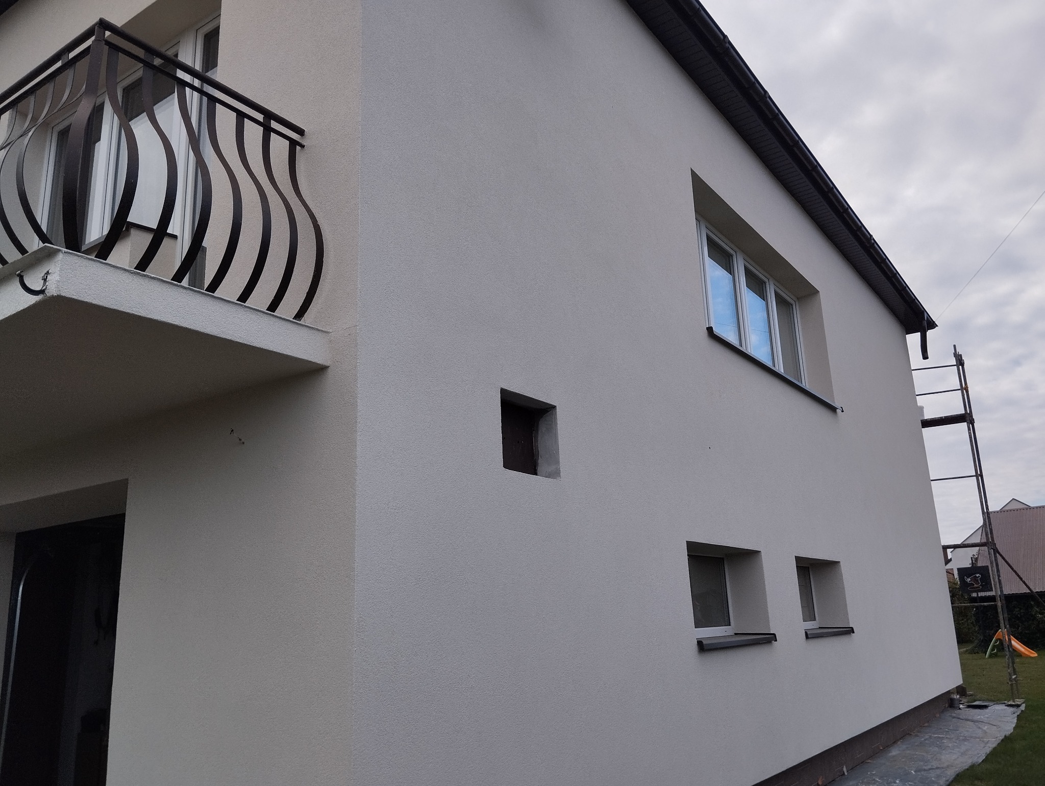 Świeżo otynkowana elewacja budynku w Przemyślu z charakterystycznym małym, kwadratowym otworem, widoczne rusztowanie i balkon z ozdobną, metalową balustradą.