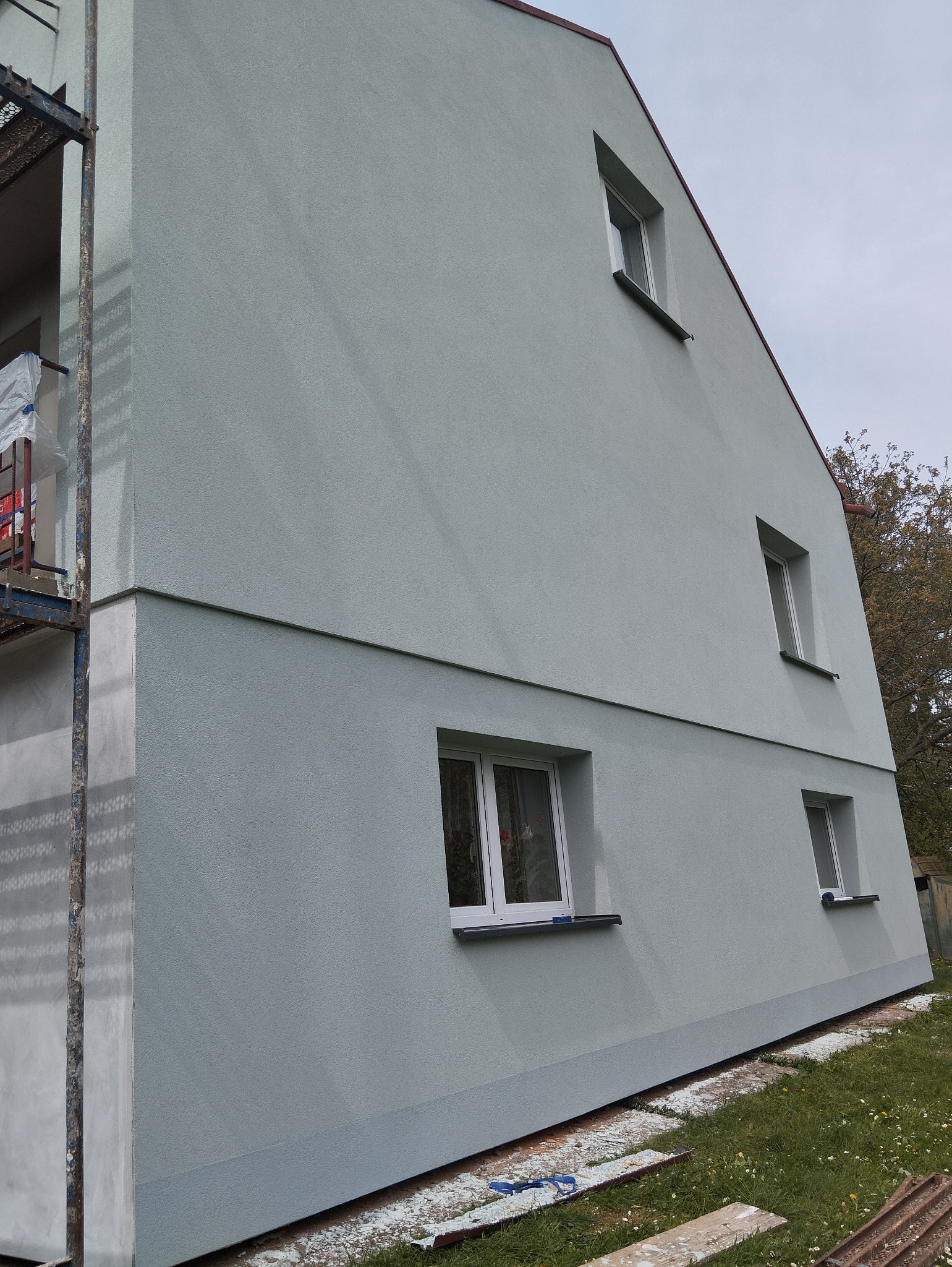 Świeżo pomalowana elewacja domu jednorodzinnego w odcieniach szarości, widoczne rusztowanie i pozostałości farby na trawie.