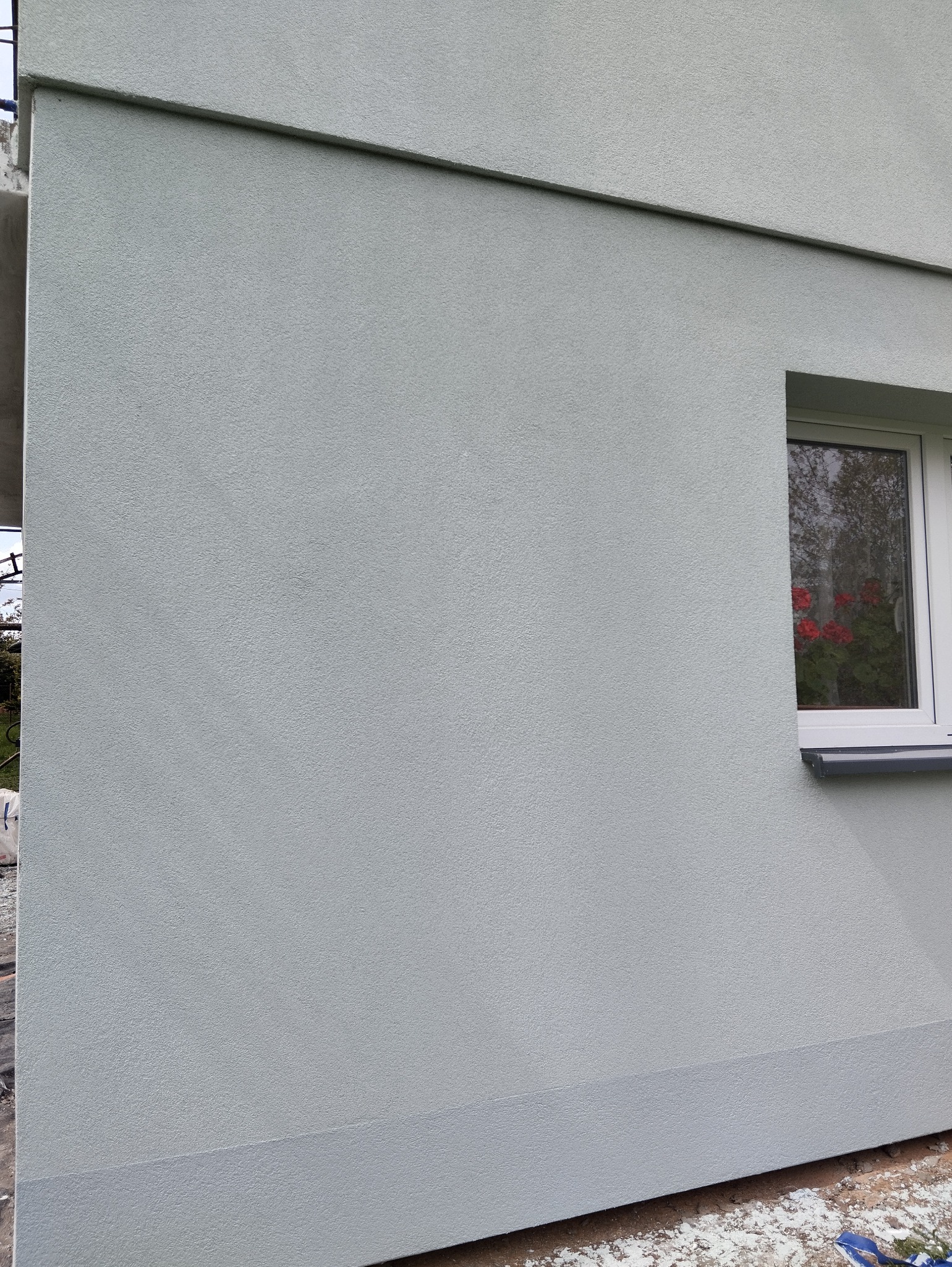 Jasnoszara elewacja budynku z widocznym oknem, na parapecie czerwone kwiaty w doniczce. Świeżo pomalowana powierzchnia z delikatną fakturą.