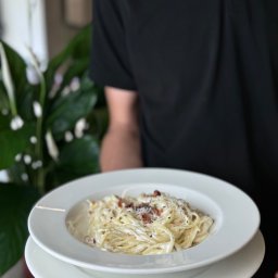 MK9 SPÓŁKA Z OGRANICZONĄ ODPOWIEDZIALNOŚCIĄ - Kelner w czarnej koszuli prezentuje talerz kremowego spaghetti carbonara na tle roślin doniczkowych.