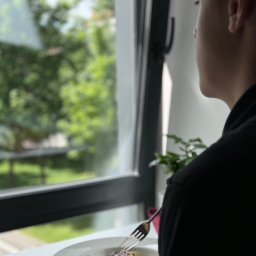 MK9 SPÓŁKA Z OGRANICZONĄ ODPOWIEDZIALNOŚCIĄ - Osoba w czarnym ubraniu je sałatkę z fioletową kapustą przy oknie z widokiem na zieleń, trzymając widelec skierowany w stronę potrawy.