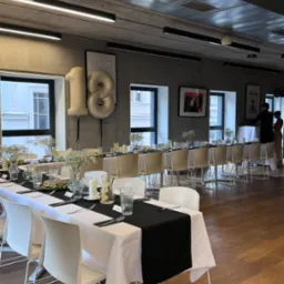 Długi stół z białym obrusem i czarnym bieżnikiem, nakryty dla wielu osób, z dekoracjami kwiatowymi i balonami w kształcie cyfr 18, przygotowany na uroczystość w nowoczesnym wnętrzu.