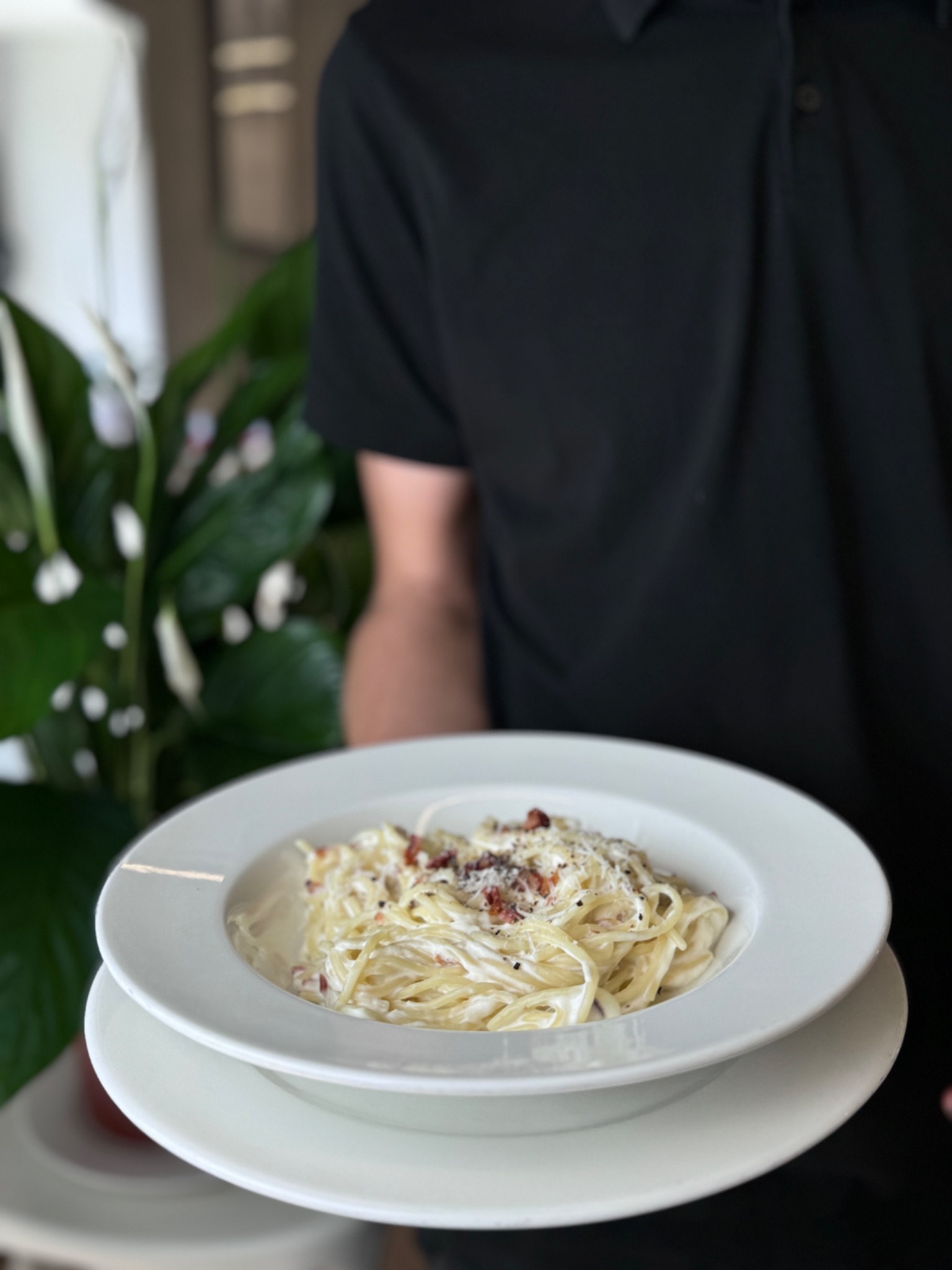 Kelner w czarnej koszuli prezentuje talerz kremowego spaghetti carbonara na tle roślin doniczkowych.