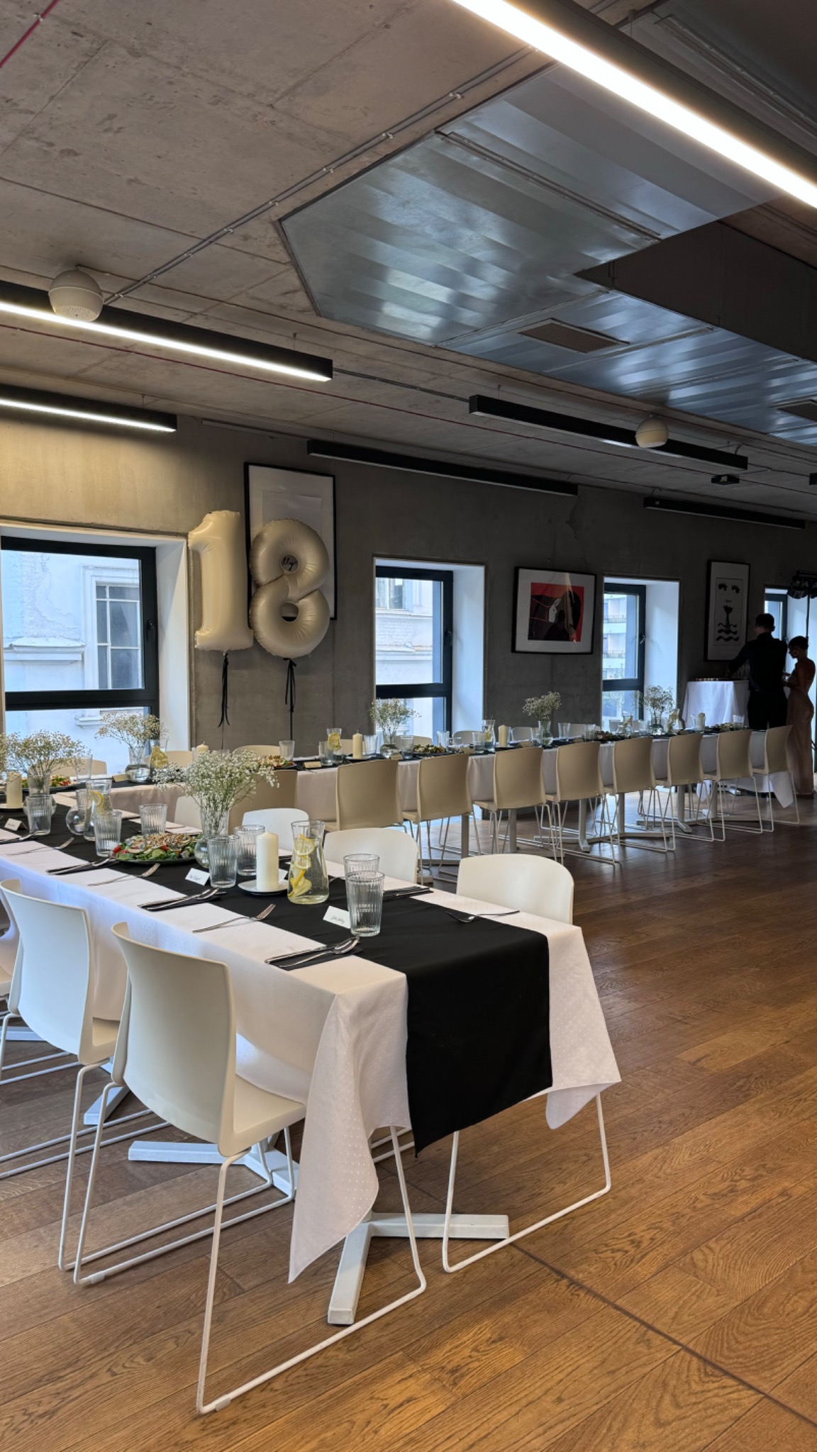 Długi stół z białym obrusem i czarnym bieżnikiem, nakryty dla wielu osób, z dekoracjami kwiatowymi i balonami w kształcie cyfr 18, przygotowany na uroczystość w nowoczesnym wnętrzu.