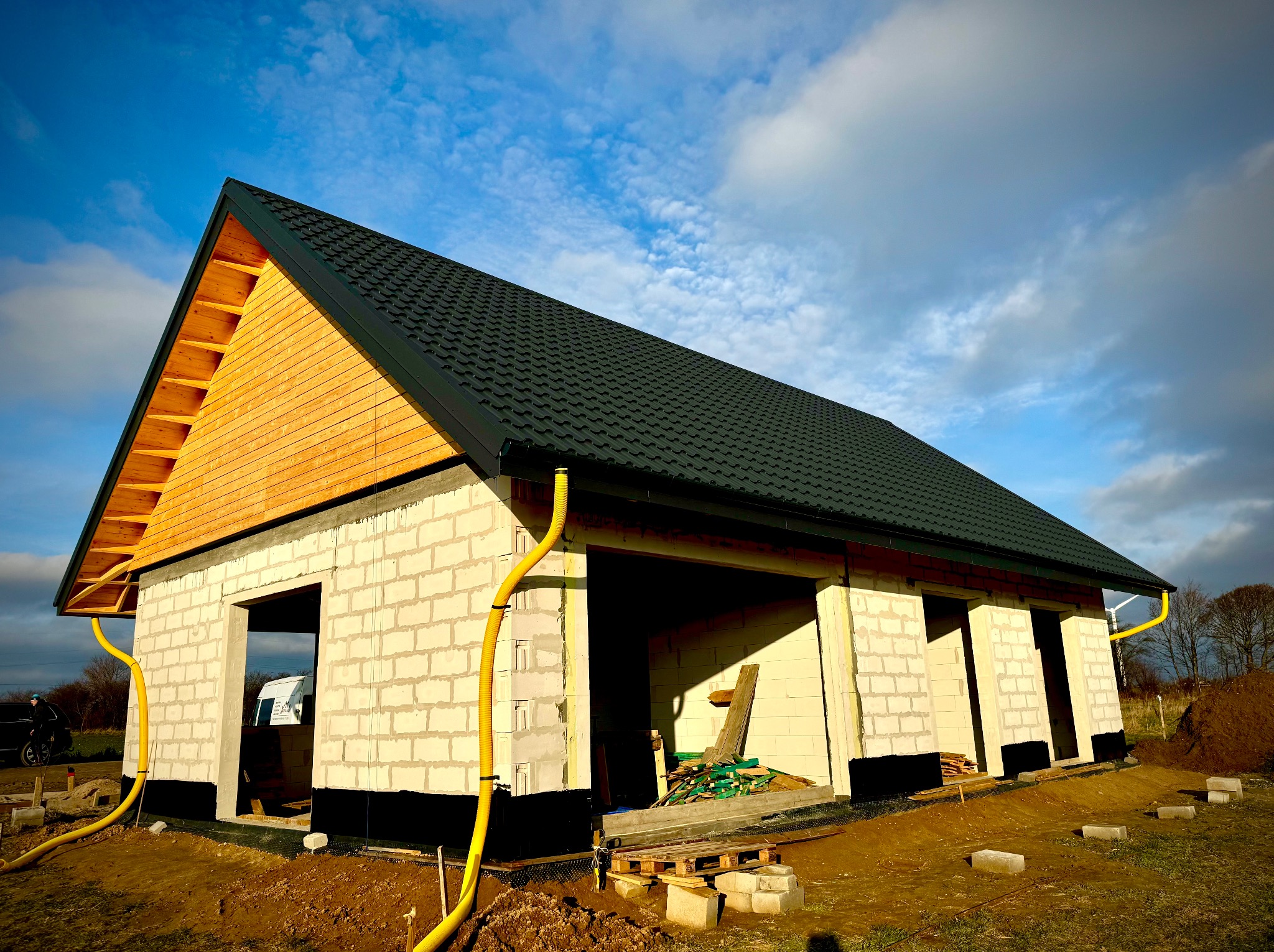 Surowy budynek w trakcie budowy z widoczną konstrukcją dachu, rynnami i ścianami z bloczków betonowych na tle błękitnego nieba z chmurami.