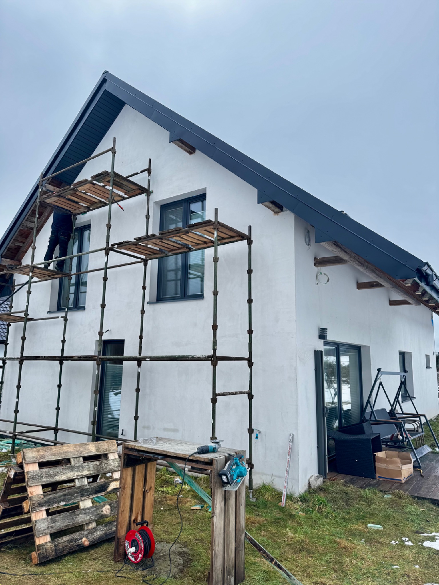 Prace dekarskie na rusztowaniu przy białym domu z ciemną podbitką dachową, widoczne narzędzia i materiały budowlane.