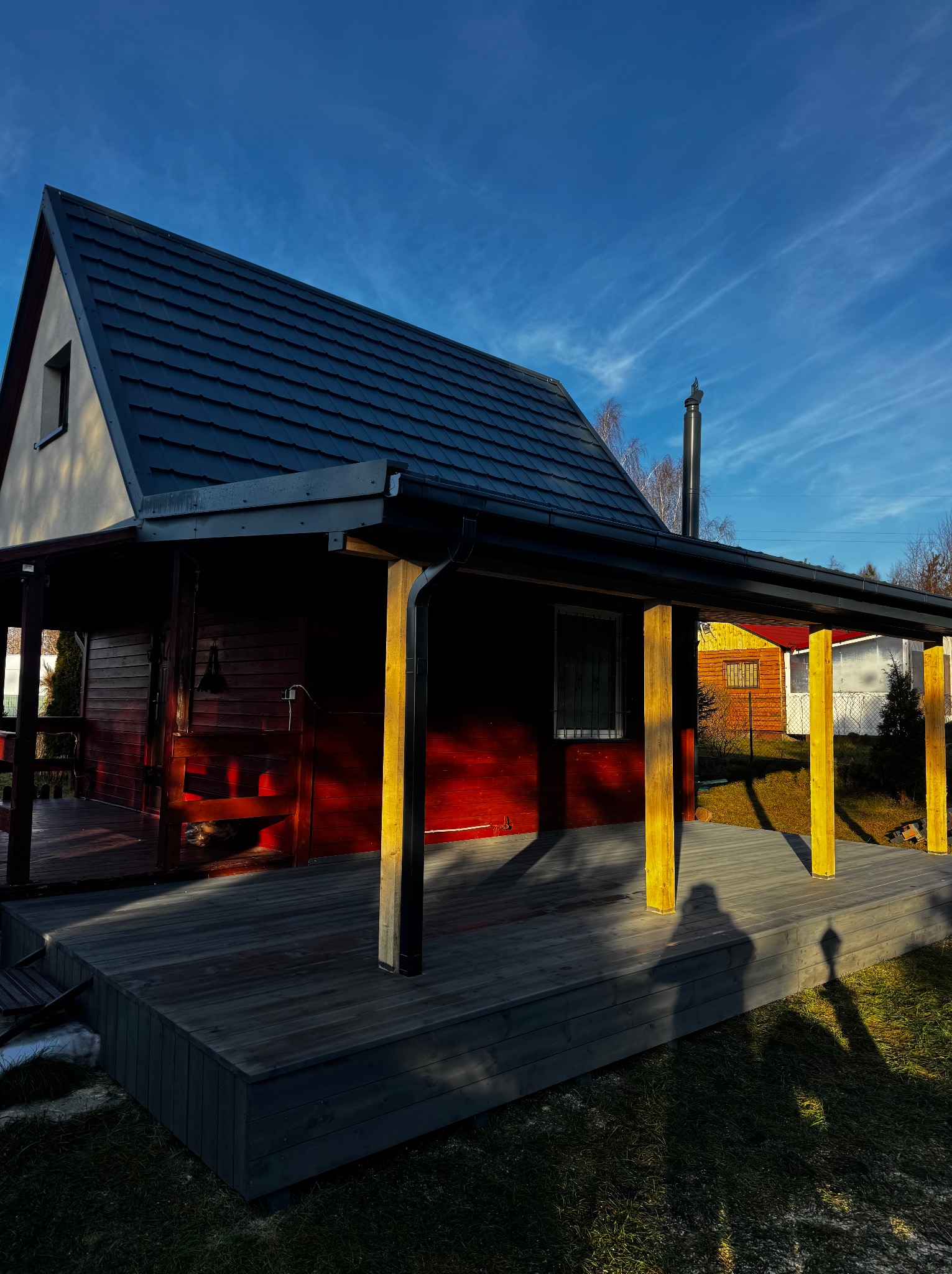 Czerwony domek z ciemnym dachem i drewnianym tarasem w promieniach zachodzącego słońca. Widoczne detale konstrukcyjne i cień osoby na trawie.