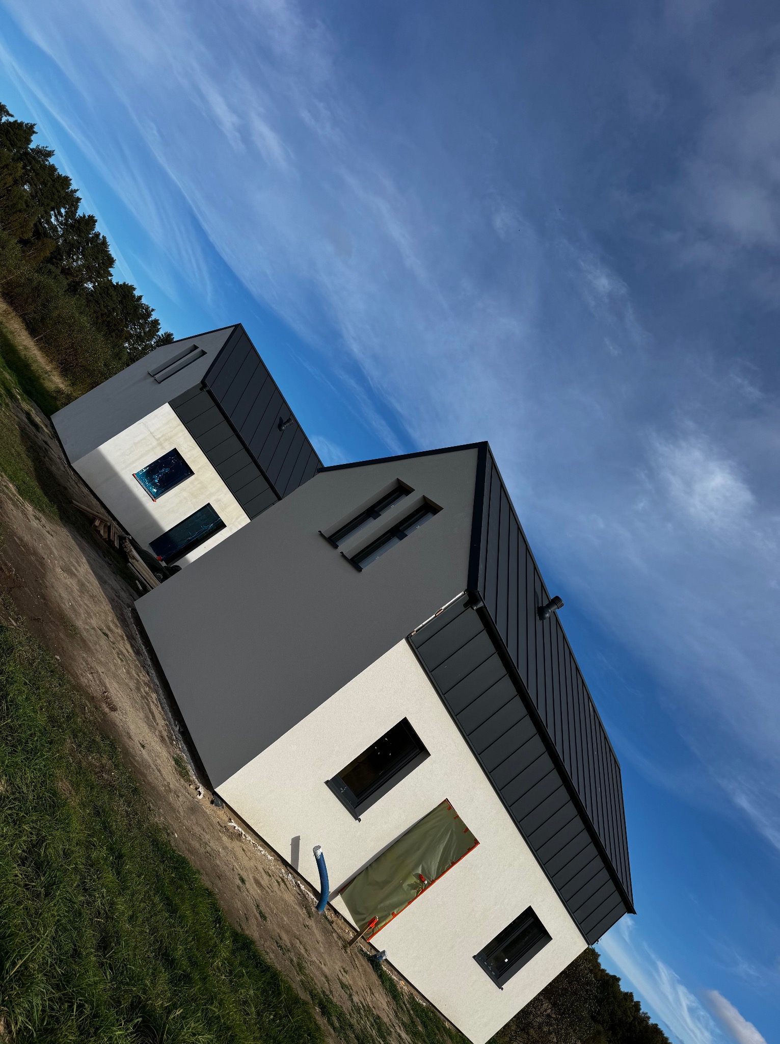 Dwa nowoczesne domy z ciemnymi dachami blachodachówką, jasną elewacją i oknami zabezpieczonymi folią, na tle błękitnego nieba z chmurami. Ujęcie pod kątem.