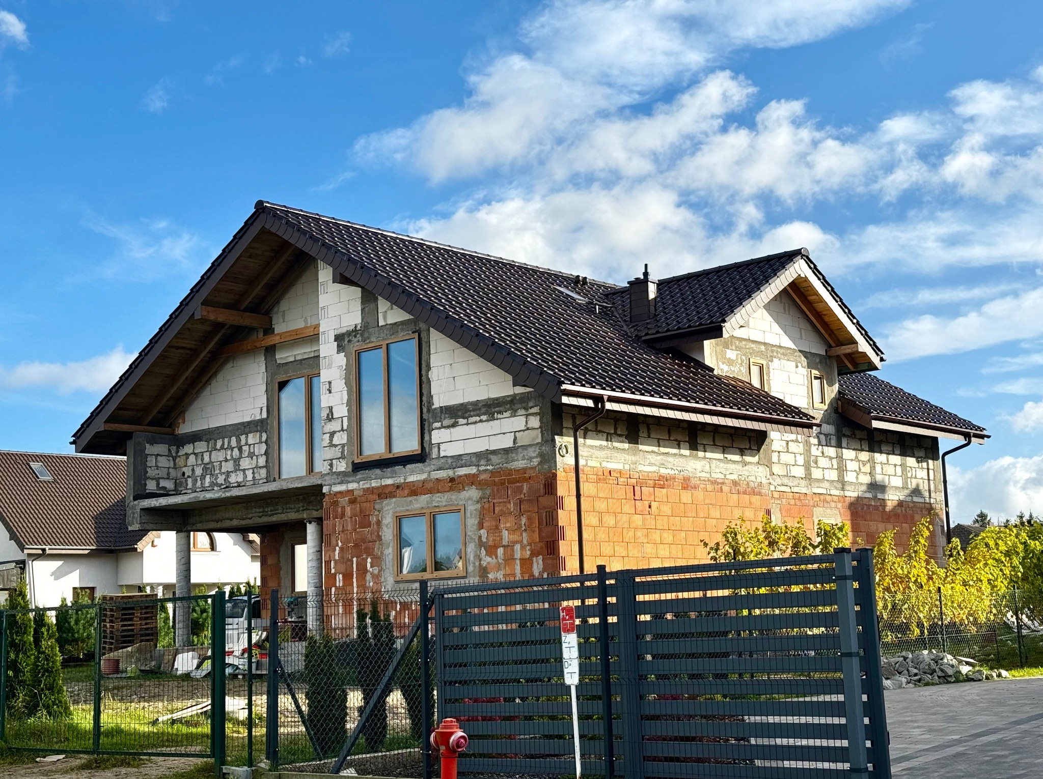 Dom w trakcie budowy z ciemnym dachem i widoczną konstrukcją ścian z pustaków, na tle błękitnego nieba z chmurami. Widoczny fragment ogrodzenia i hydrant.