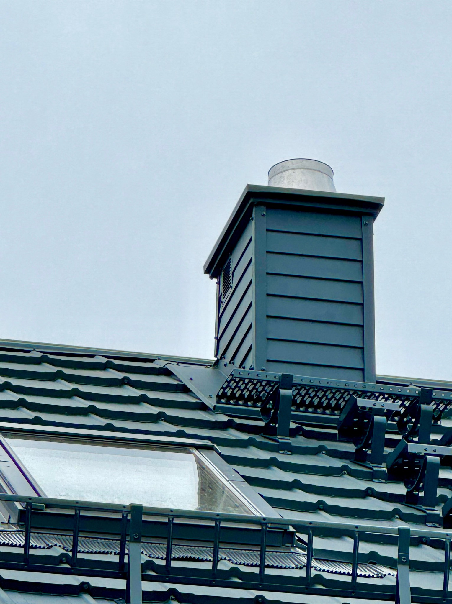 Grafitowy dach z oknem połaciowym i kominem wentylacyjnym, zabezpieczony barierkami śniegowymi. Ujęcie z dołu, pochmurne niebo w tle.