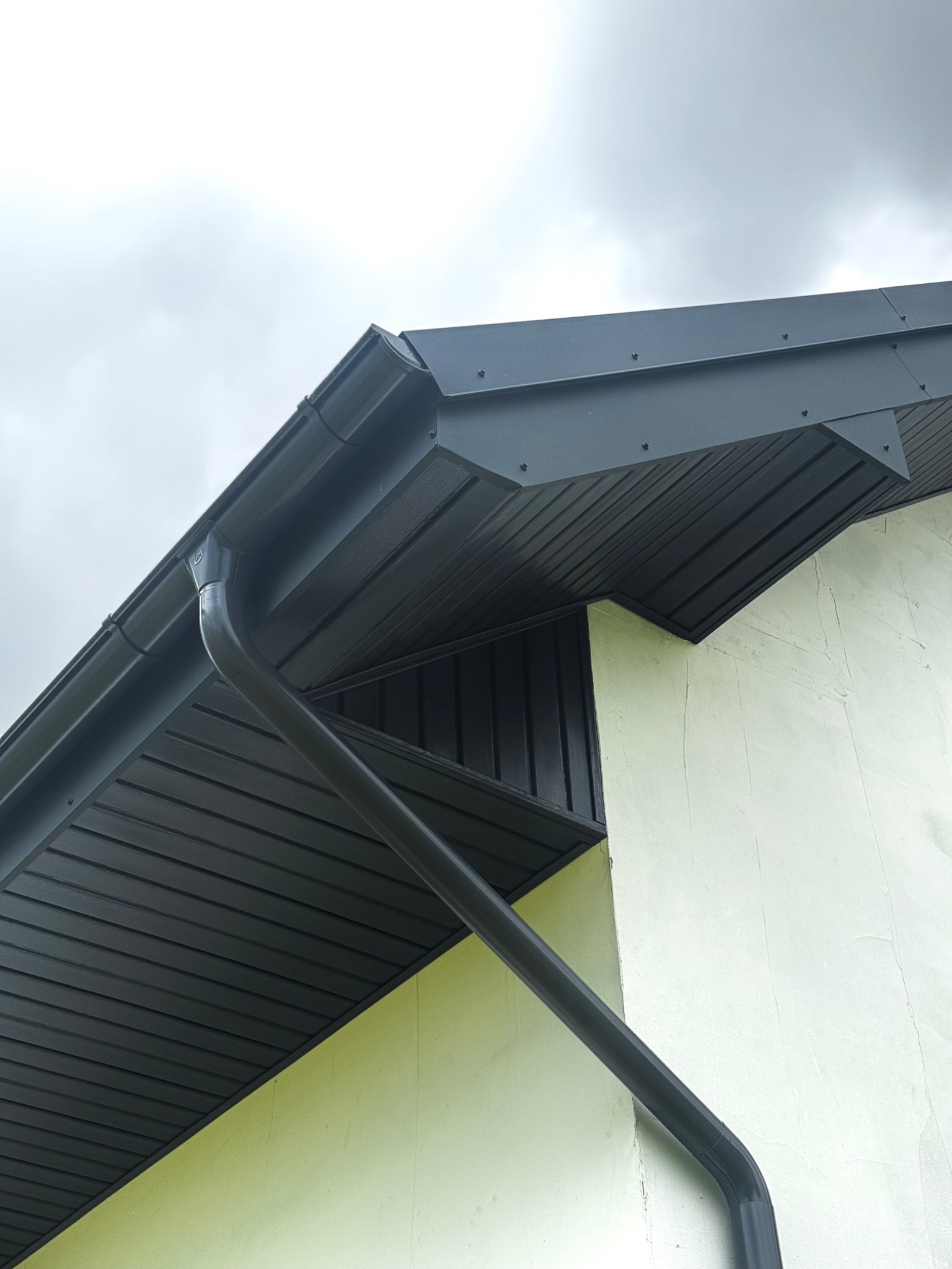 Grafitowa rynna spustowa i podbitka dachowa z widocznymi mocowaniami na tle pochmurnego nieba i fragmentu ściany budynku o jasnej elewacji.