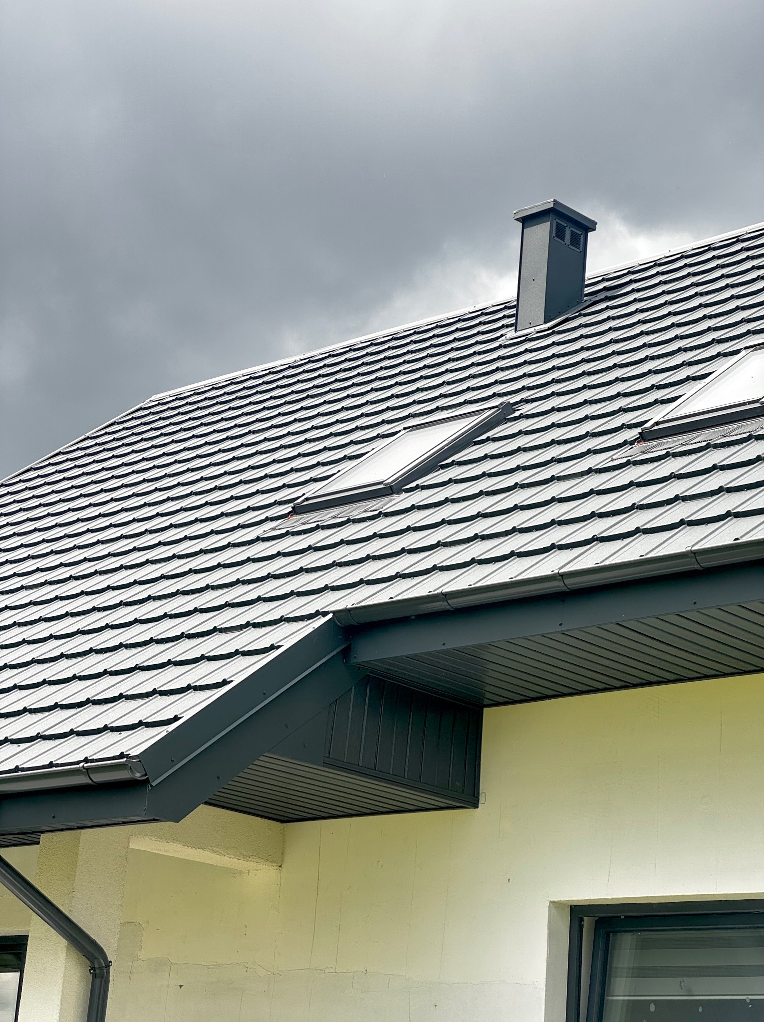 Szary dach z dachówką karpiówką i oknami połaciowymi na tle pochmurnego nieba. Widoczny komin i fragment ściany budynku z rynną.