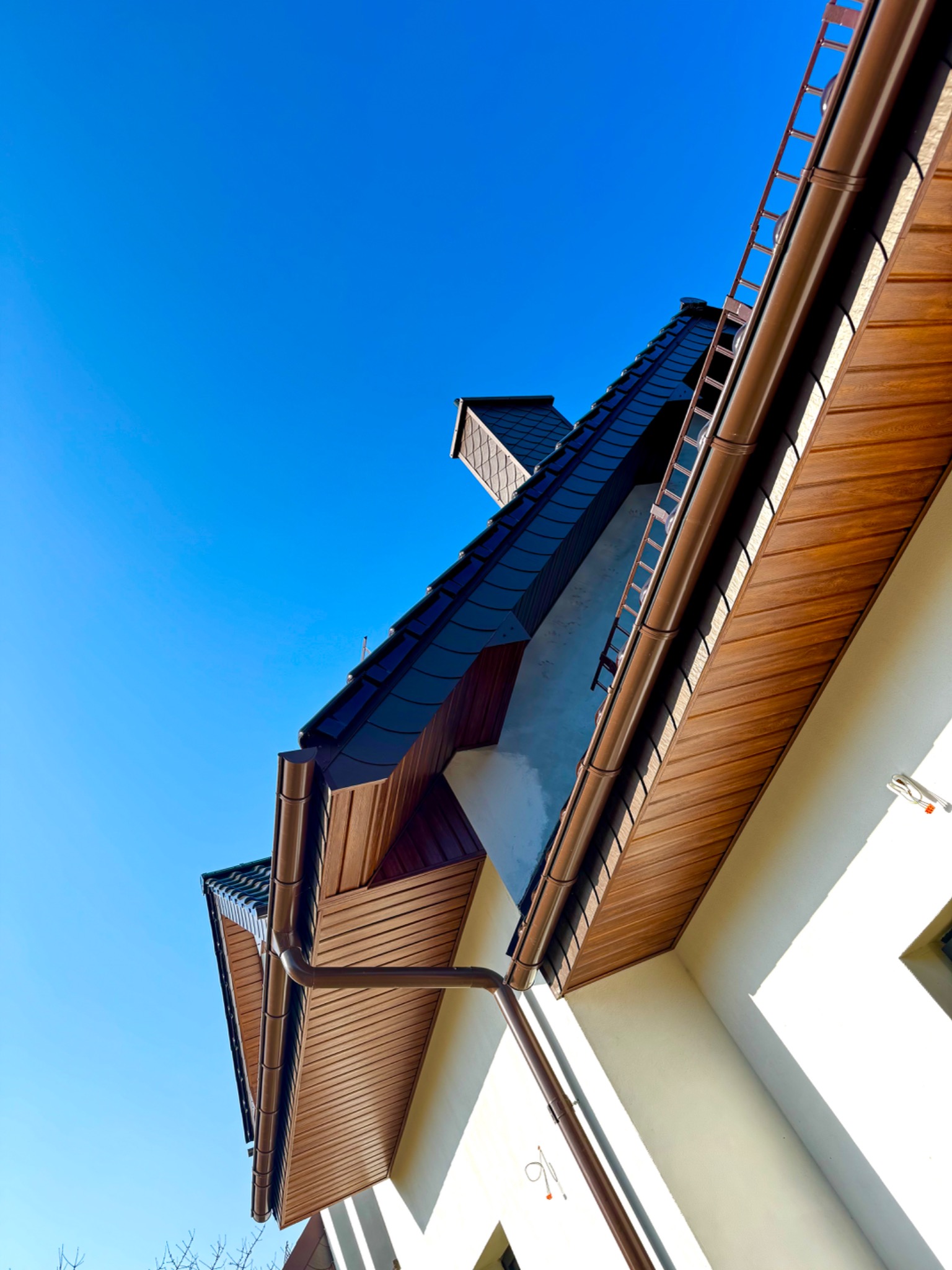 Ukończony dach z brązowymi rynnami i podbitką, widziany pod kątem z dołu na tle błękitnego nieba. Widoczny fragment elewacji budynku.