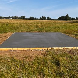 RAFAŁ MICKA RAFBRUK - Wylana płyta betonowa fundamentowa otoczona drewnianym szalunkiem na zewnątrz, na tle łąki i lasu, w słoneczny dzień.