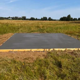 Wylana płyta betonowa fundamentowa otoczona drewnianym szalunkiem na zewnątrz, na tle łąki i lasu, w słoneczny dzień.