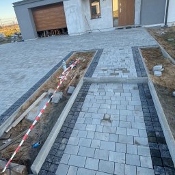 RAFAŁ MICKA RAFBRUK - Układanie szarej kostki brukowej z ciemnoszarym obramowaniem, częściowo zakończone, z wykopanymi miejscami na rabaty kwiatowe zabezpieczonymi taśmą ostrzegawczą, zagęszczarka w tle.