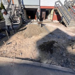 RAFAŁ MICKA RAFBRUK - Prace przygotowawcze do ułożenia nawierzchni z kostki: wyrównywanie terenu, widoczna warstwa piasku, stos kostki granitowej i dwóch pracowników w tle.