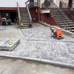 RAFAŁ MICKA RAFBRUK - Układanie szarej kostki brukowej na podjeździe, widoczna zagęszczarka wibroprasa, paleta z kostką i żółte rękawice robocze, w tle schody zewnętrzne i garaż.
