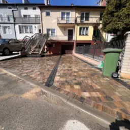 Ułożona kostka brukowa w kolorach ziemi tworzy wzorzysty podjazd do garażu. Widoczne domy w tle i samochód. Na pierwszym planie krawężnik oddzielający kostkę od ulicy.