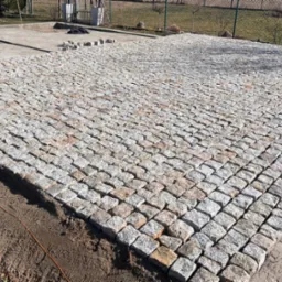 Ułożona kostka granitowa w regularny wzór, widoczne fragmenty terenu przed i za. Częściowo ułożona nawierzchnia z jasnoszarej kostki.