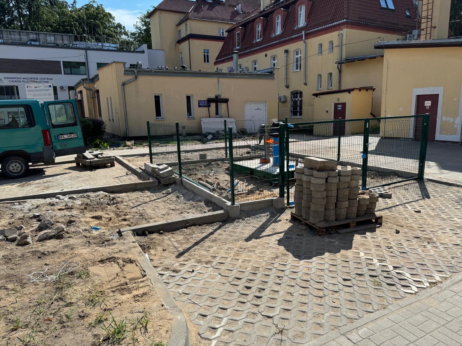Przygotowanie terenu pod nawierzchnię z betonowej kostki brukowej, widoczne obrzeża, paleta z kostką, częściowo ułożona nawierzchnia, teren budowy zabezpieczony ogrodzeniem, budynek w tle.