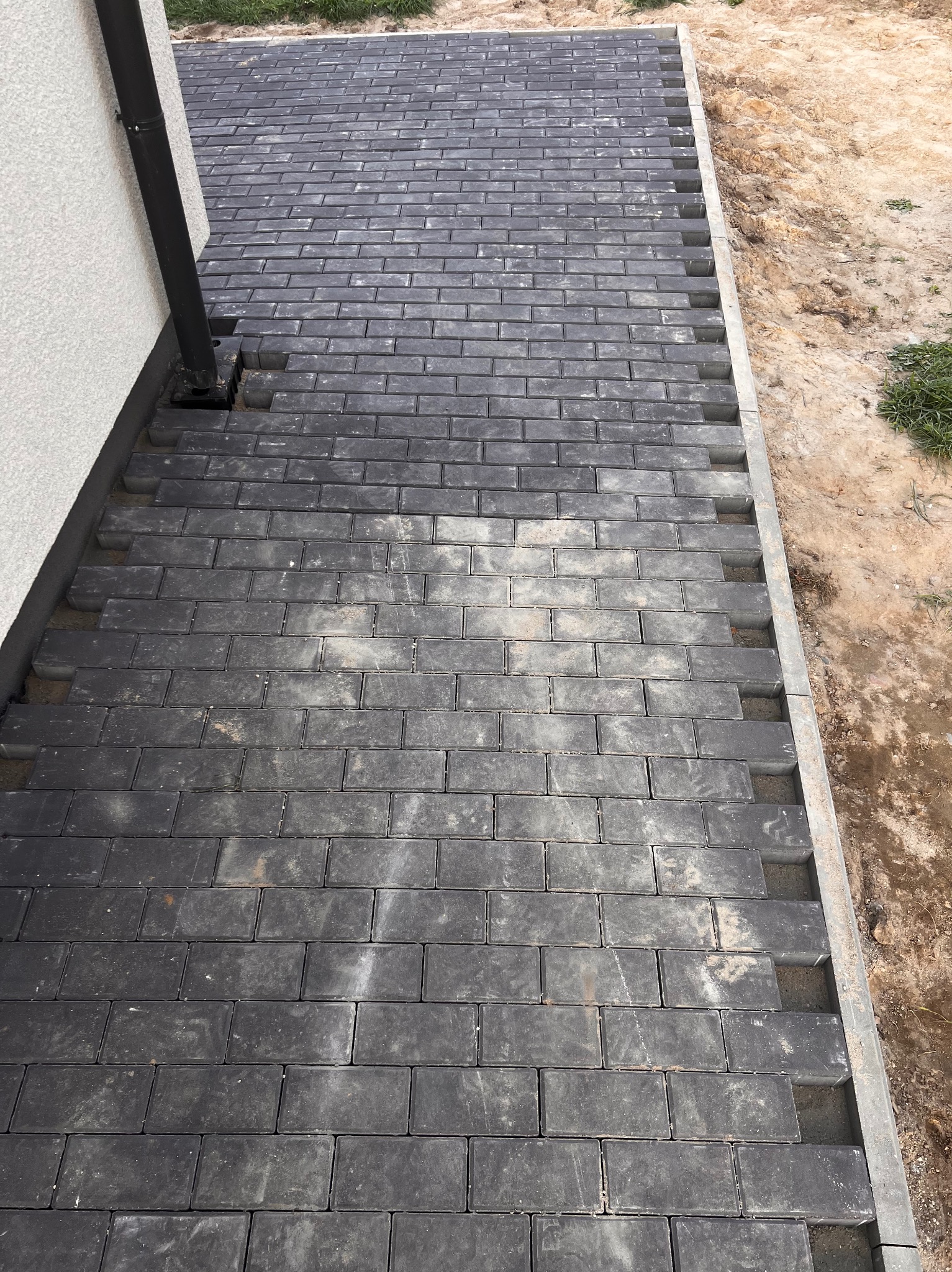 Ciemnoszara kostka brukowa ułożona przy ścianie budynku i rynnie, widoczne niedokończone krawędzie i piasek.