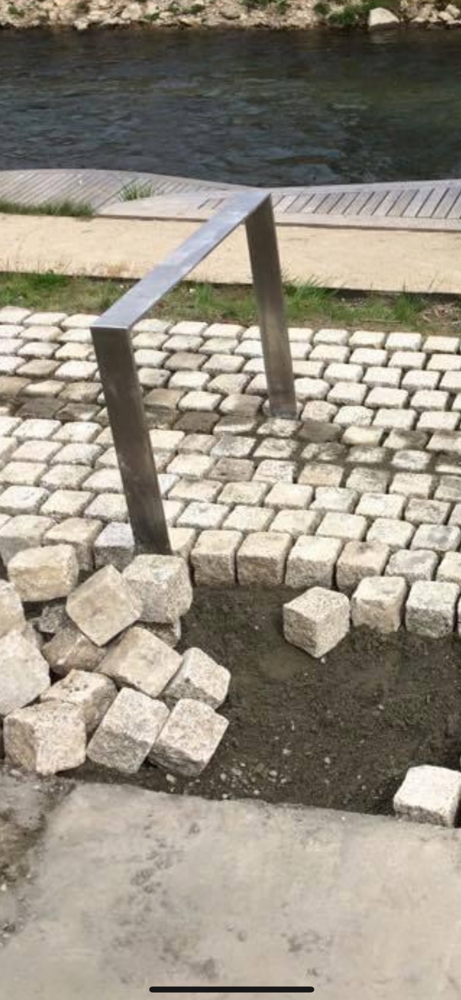 Fragment chodnika z kostki granitowej w trakcie budowy, obok rzeki, z metalowym słupkiem. Część kostek jeszcze nieułożona, leżąca obok wykopu.