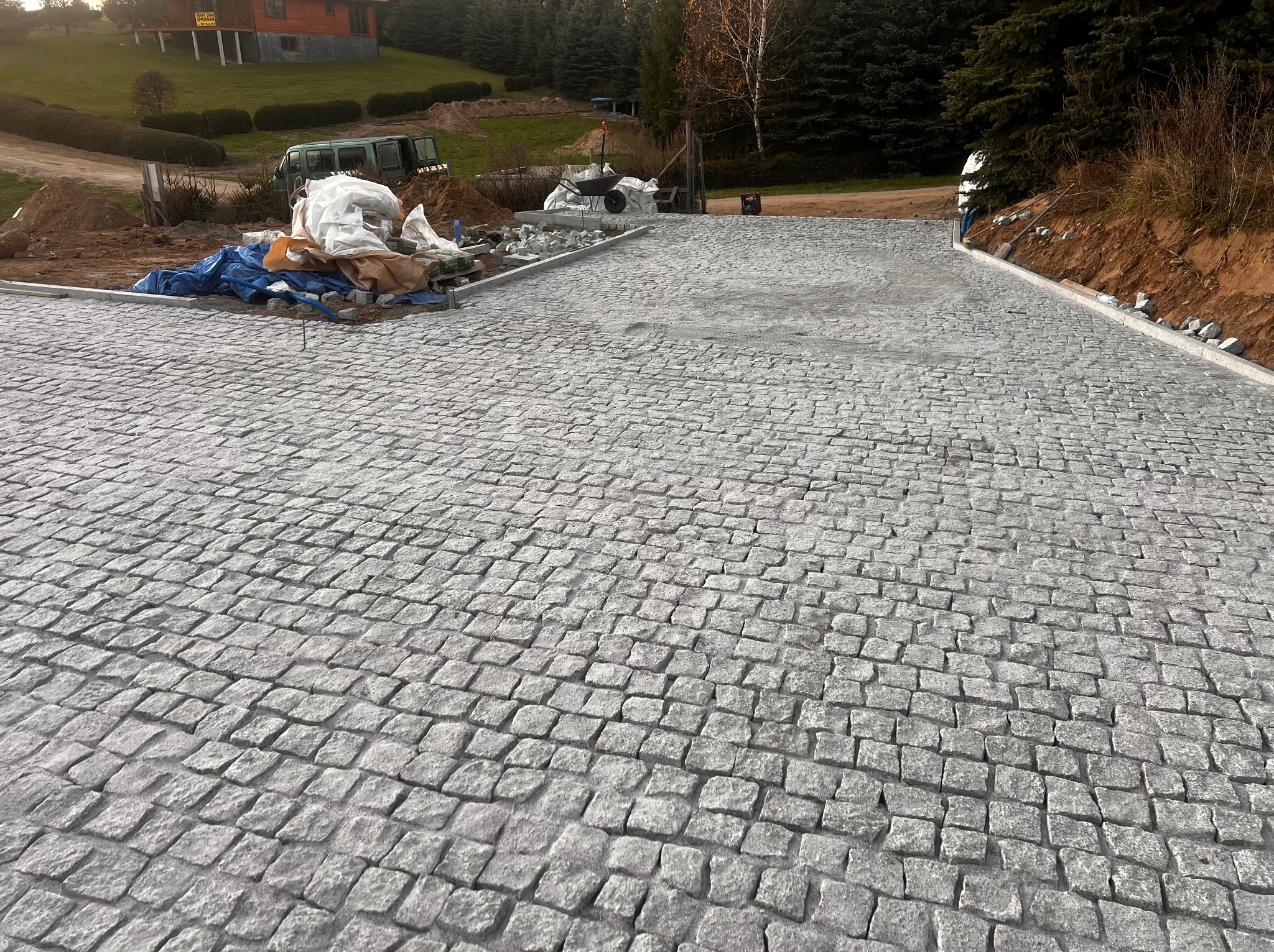 Ułożona szara kostka granitowa tworząca podjazd, z widocznymi materiałami budowlanymi i narzędziami w tle, częściowo zasłoniętymi plandeką, oraz budynkiem mieszkalnym w oddali.