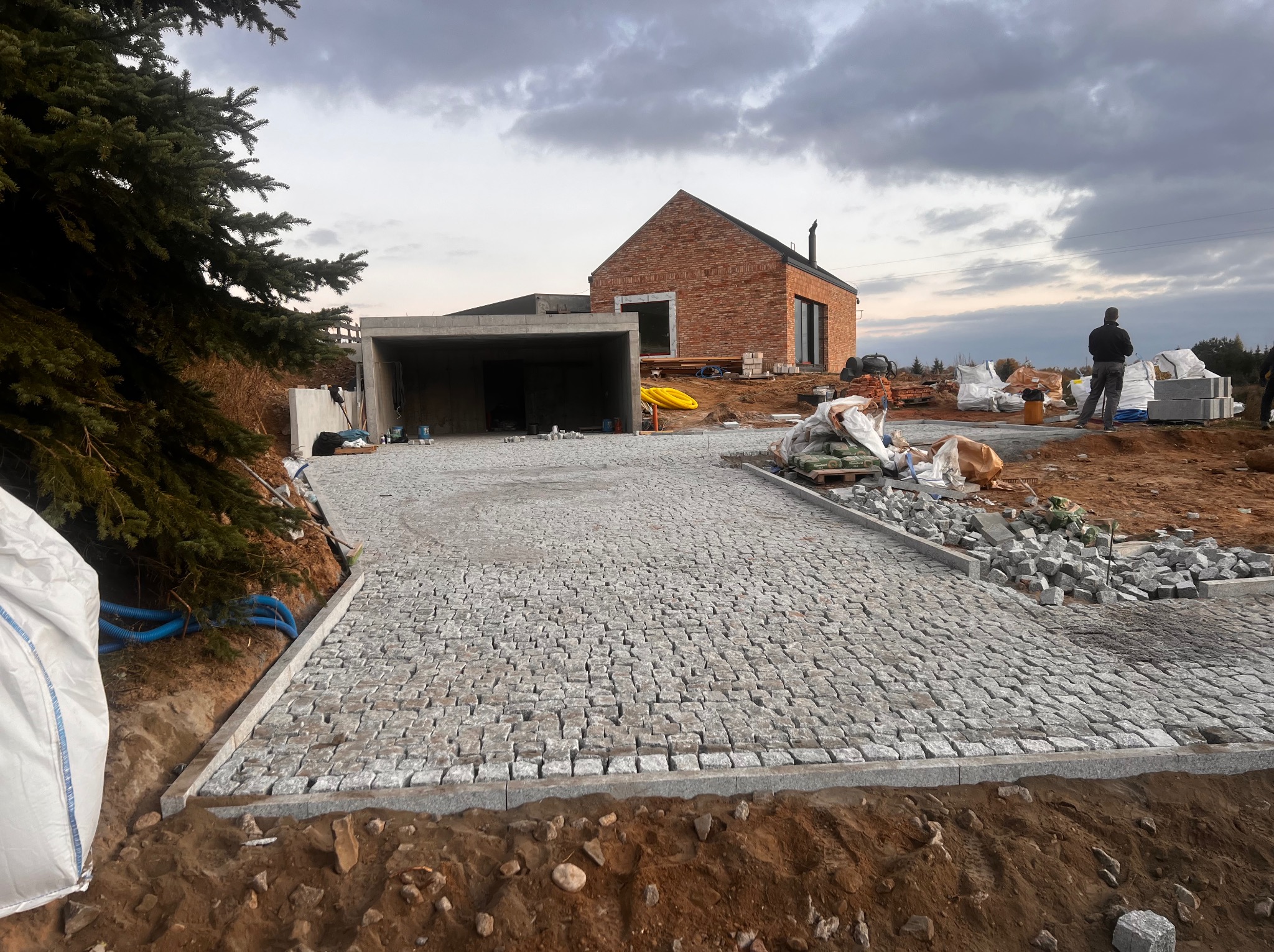 Nowa nawierzchnia z kostki granitowej na podjeździe do domu z garażem, widoczne worki z materiałami budowlanymi i osoba w tle, pochmurne niebo.