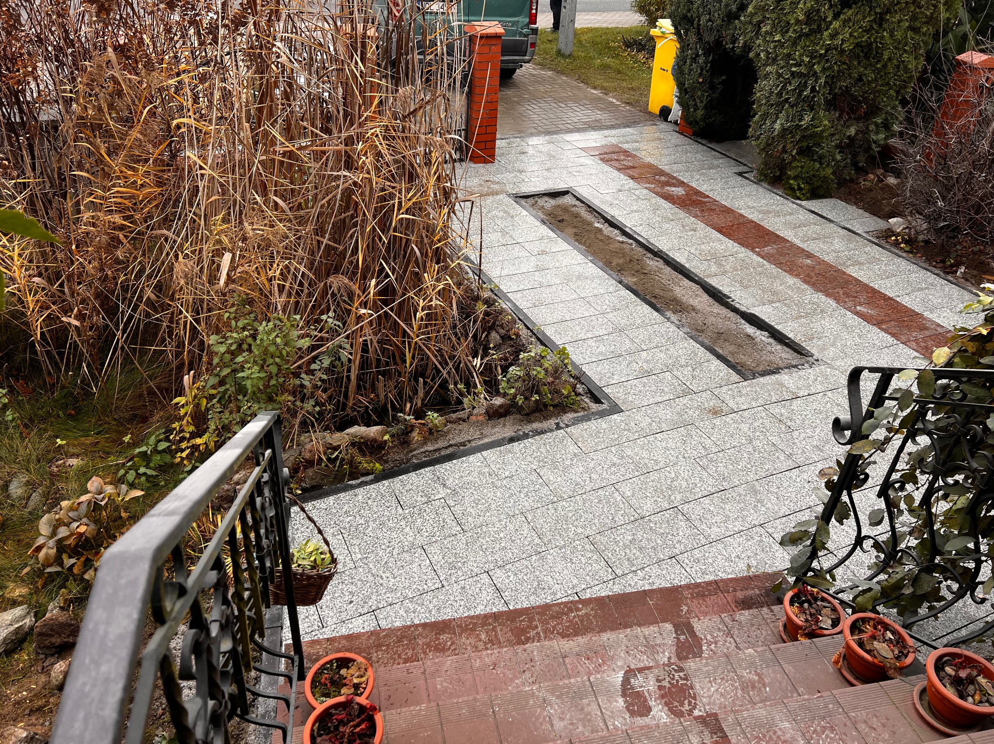 Ułożona szara kostka brukowa z dwoma wąskimi, wydłużonymi zagłębieniami wypełnionymi ziemią, obok rabaty z suchą trawą, widok z góry schodów z doniczkami.