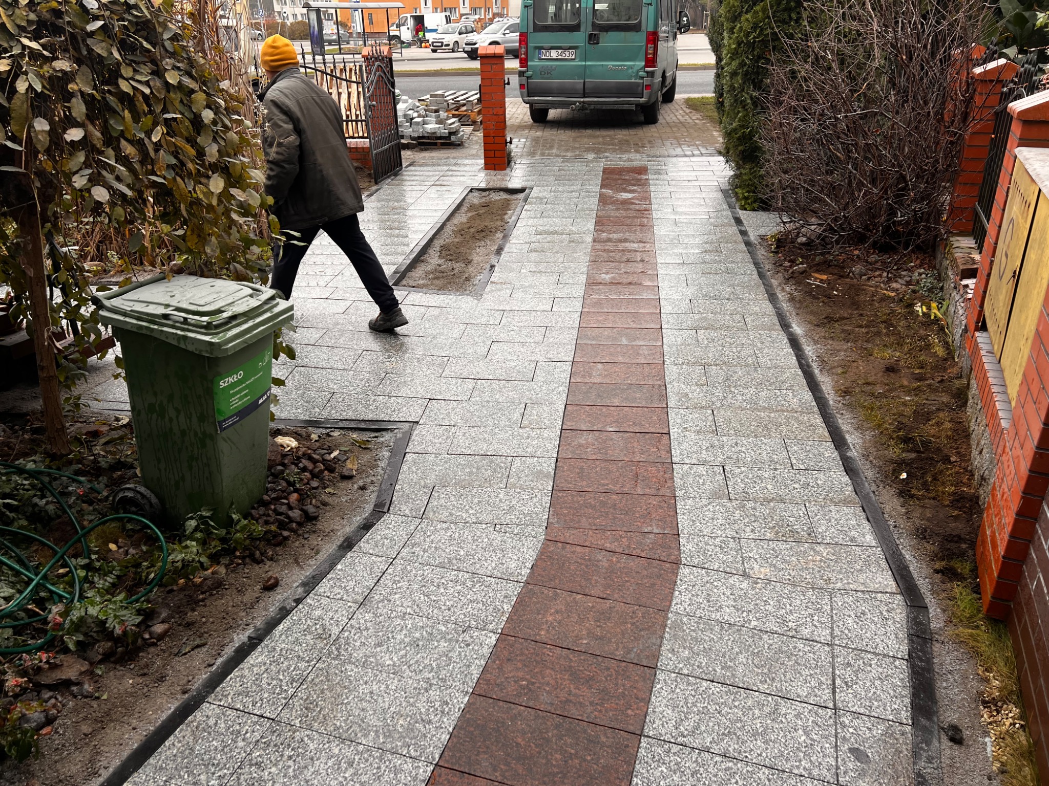 Ułożona ścieżka z kostki granitowej w dwóch kolorach, z widocznym fragmentem przygotowanego podłoża pod rośliny, na tle ulicy i zaparkowanego samochodu dostawczego, z pracownikiem w żółtej czapce...