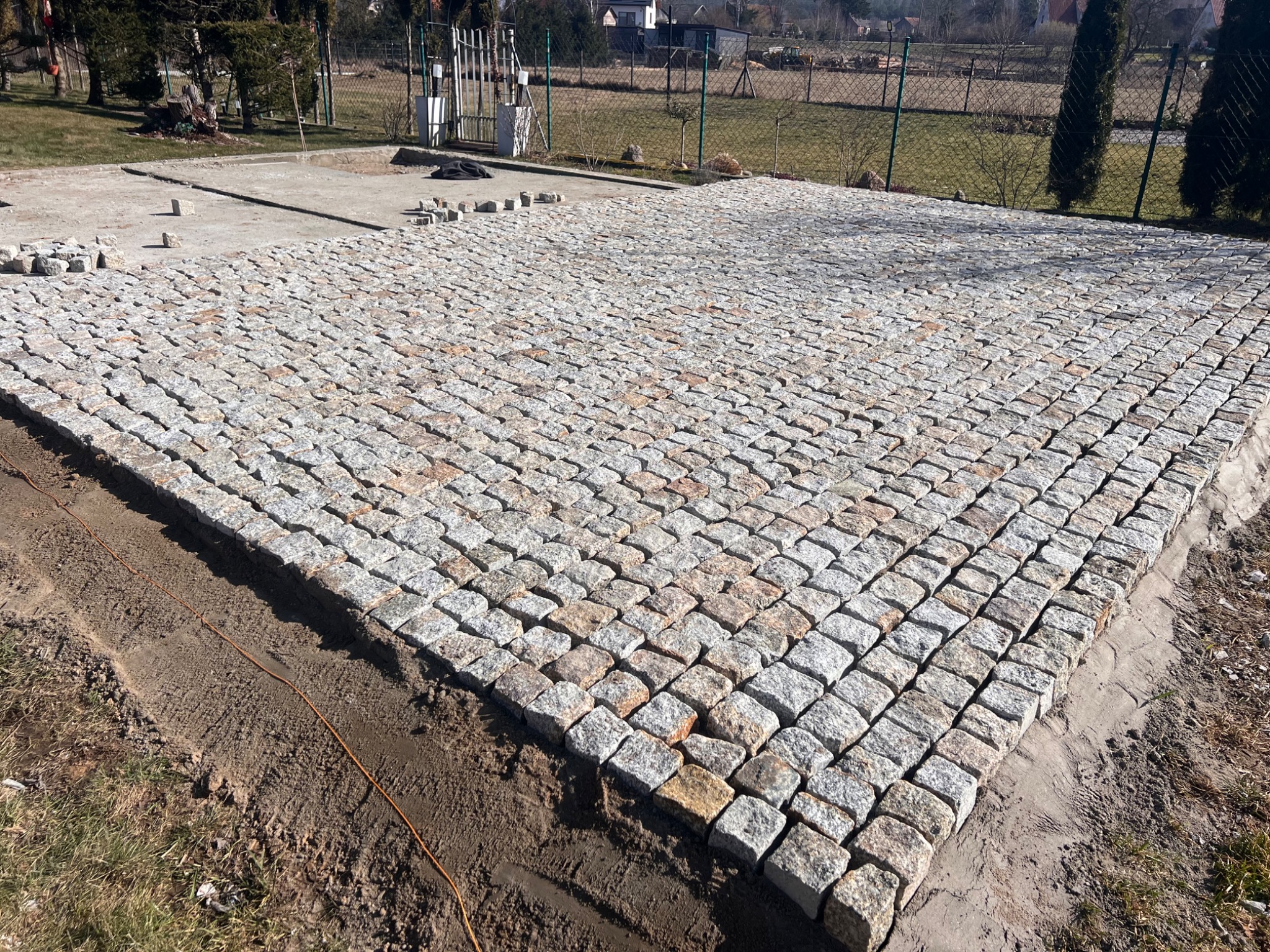 Powierzchnia z kostki granitowej w trakcie układania, widoczne przygotowane podłoże i zapas kostek, w tle ogrodzenie posesji.