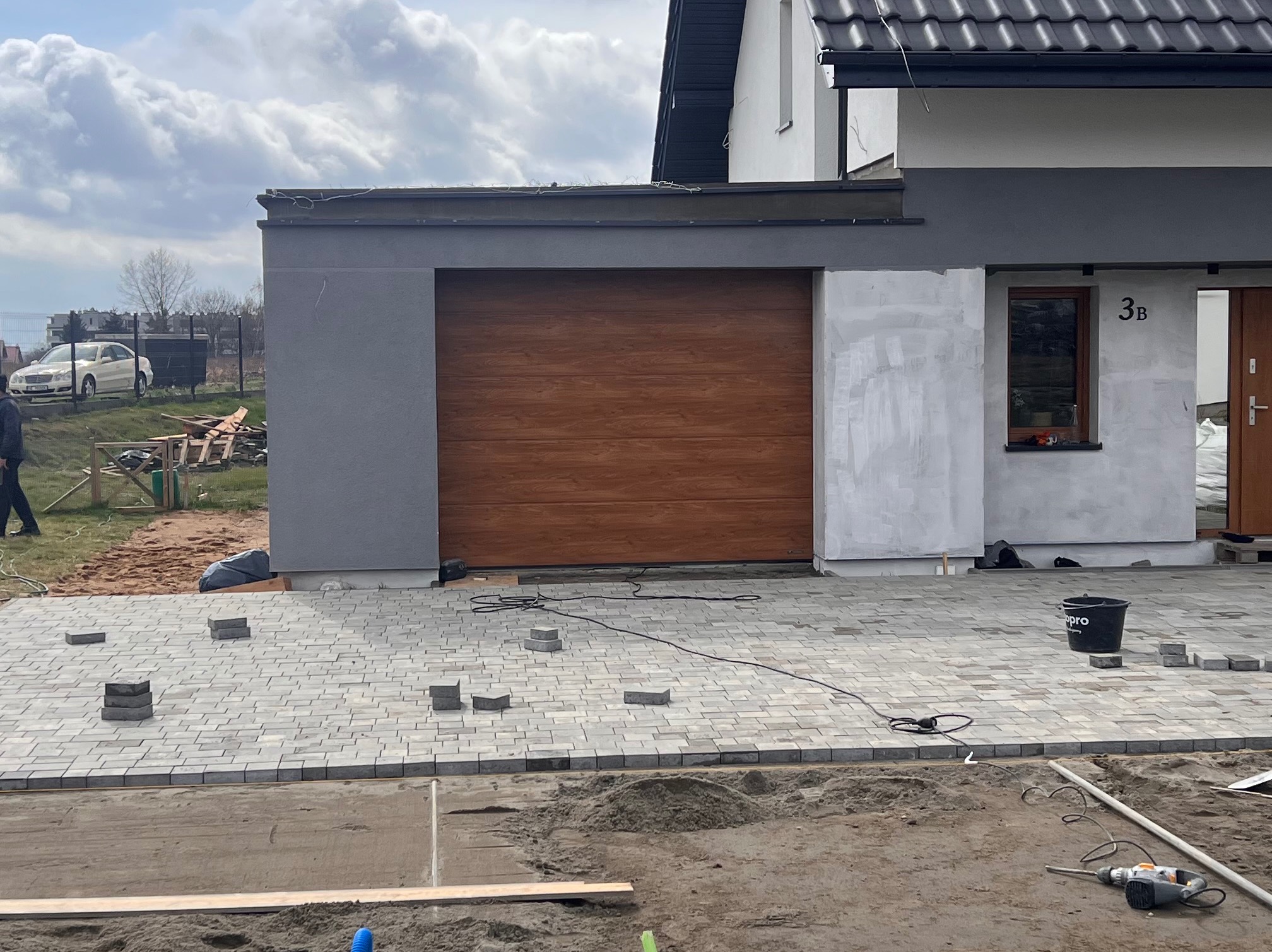 Kładzenie szarej kostki brukowej na podjeździe przed domem z brązową bramą garażową i numerem 3B, widoczne narzędzia i wiadro.