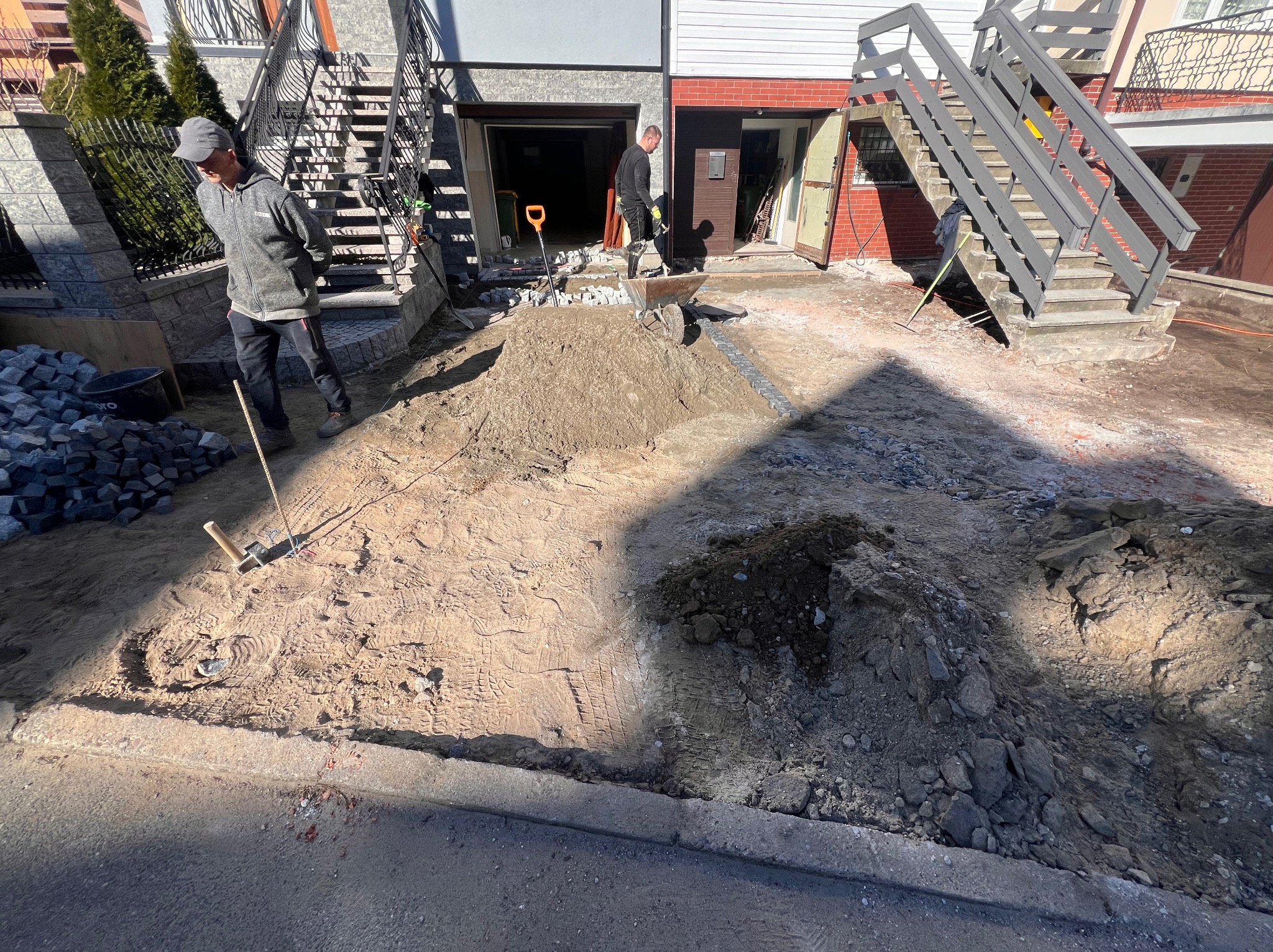 Prace przygotowawcze do ułożenia nawierzchni z kostki: wyrównywanie terenu, widoczna warstwa piasku, stos kostki granitowej i dwóch pracowników w tle.