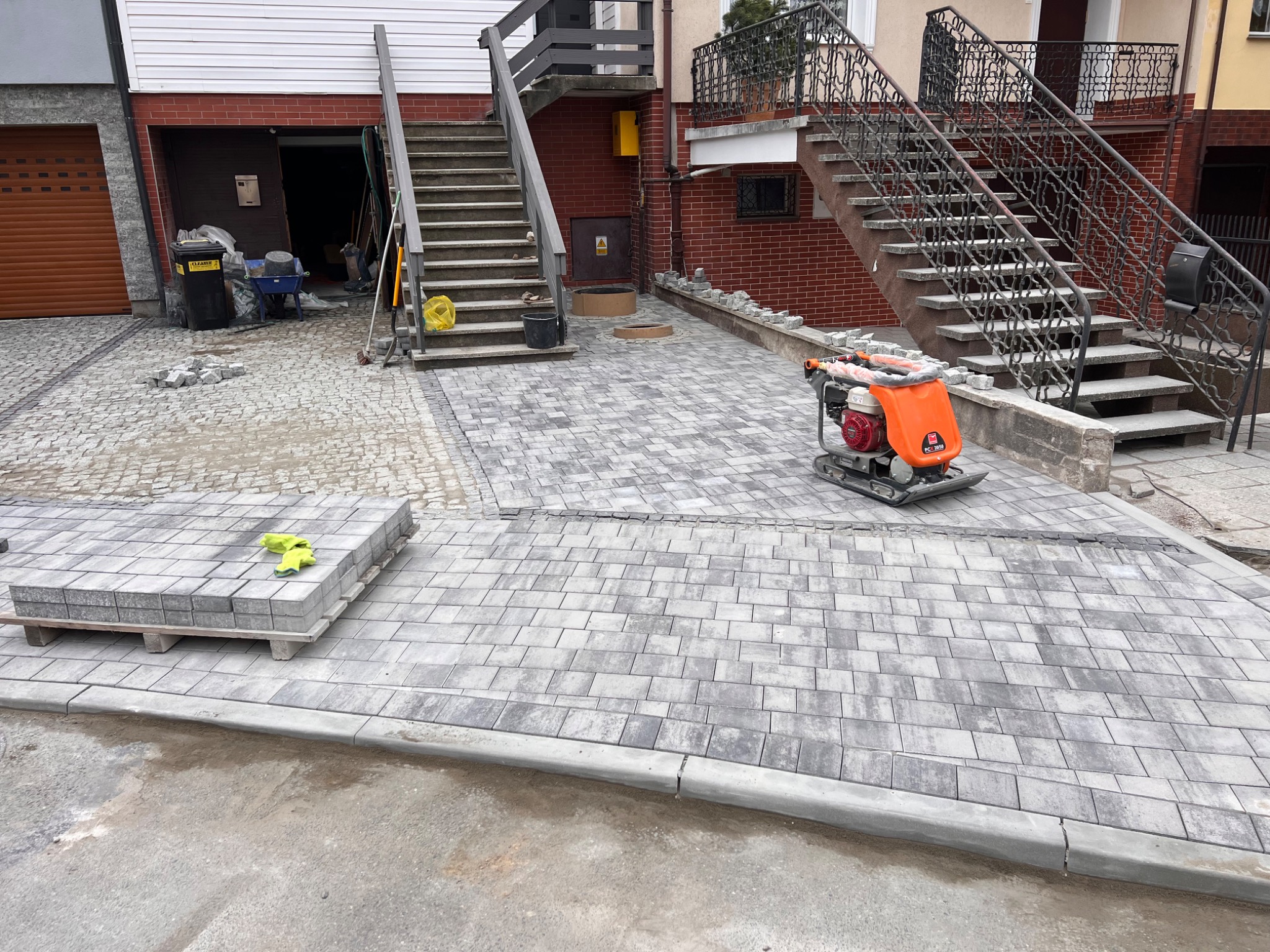 Układanie szarej kostki brukowej na podjeździe, widoczna zagęszczarka wibroprasa, paleta z kostką i żółte rękawice robocze, w tle schody zewnętrzne i garaż.