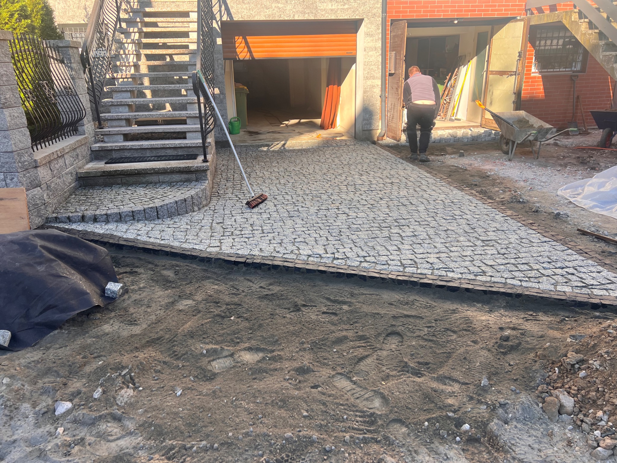 Fragment podjazdu z kostki granitowej w trakcie układania, widoczny pracownik, przygotowane podłoże, schody z kamienia i garaż w tle.